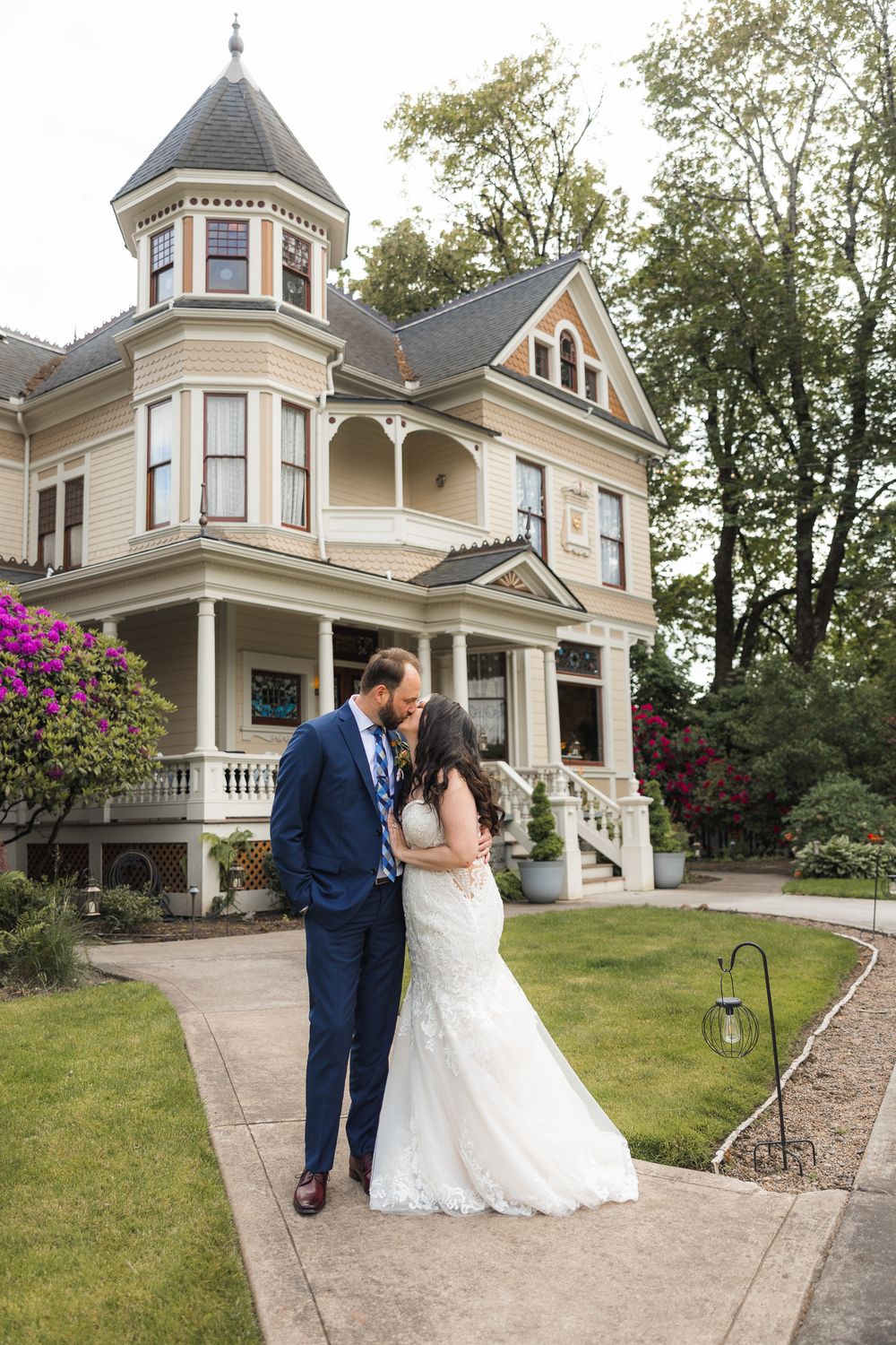 Romantic wedding couple poses outside a beautiful Victorian mansion with white columns and wrap-around porch.