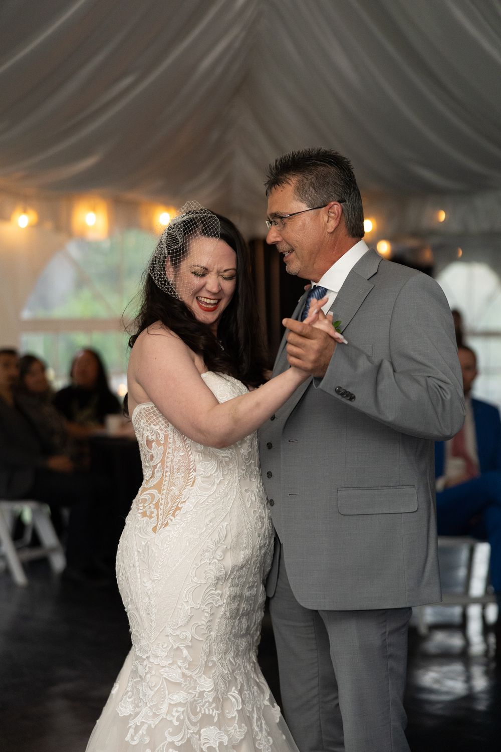 A couple dances together under a white tent reception, with the bride wearing a lace gown.
