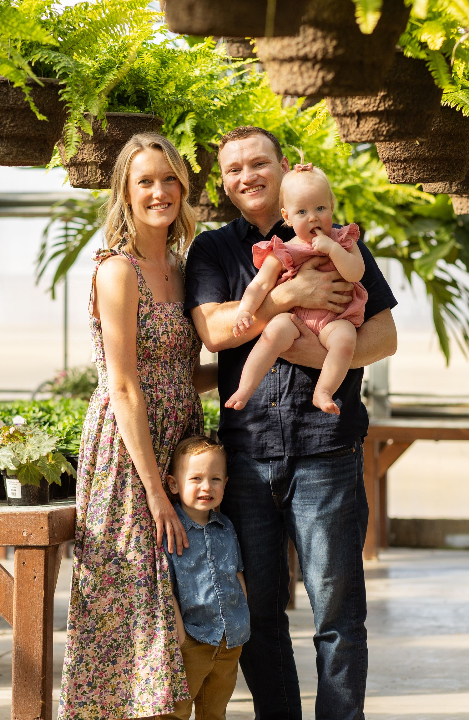 Happy family portrait in a greenhouse surrounded by hanging ferns and natural light.