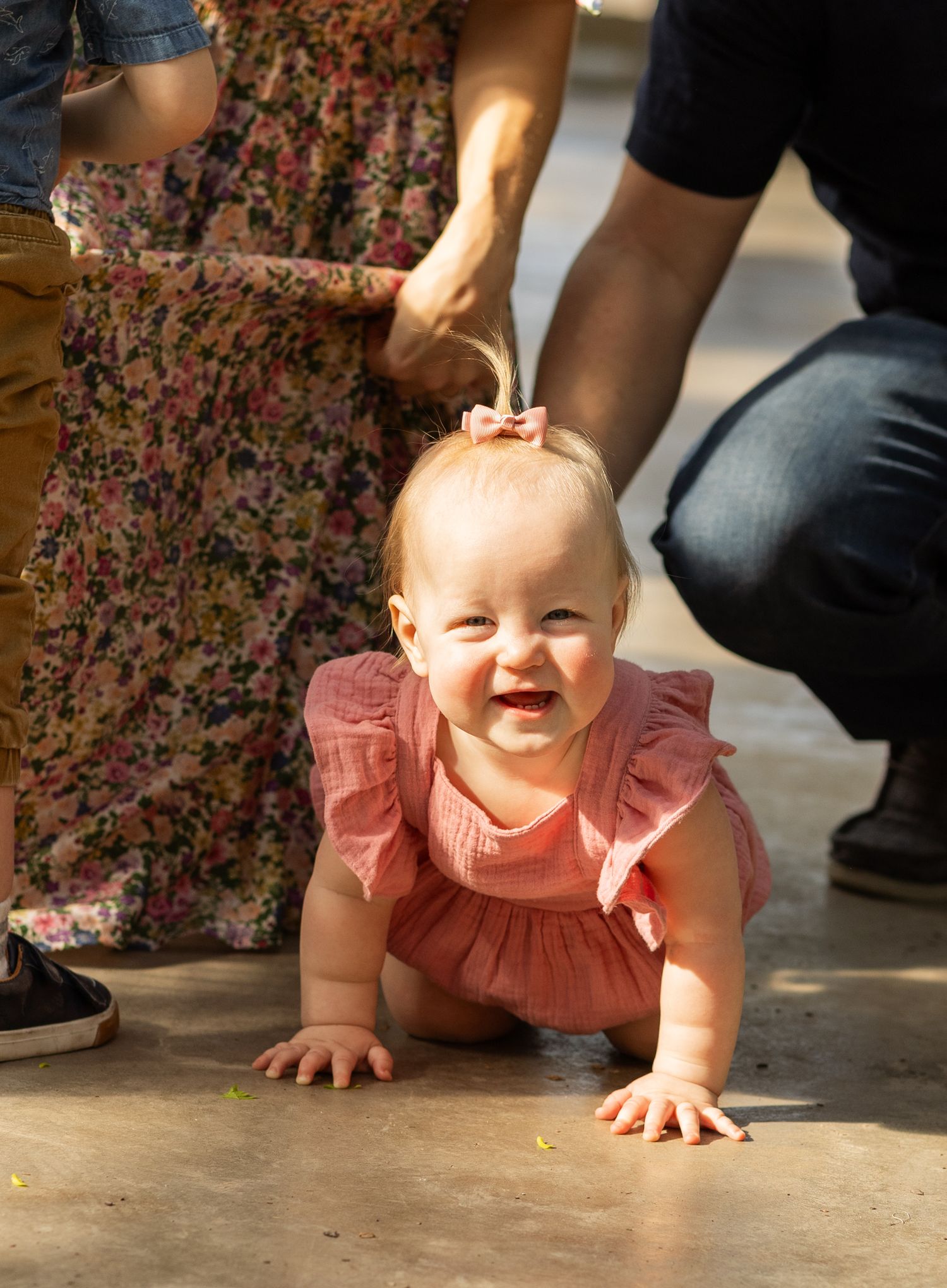 A smiling baby in a pink ruffled dress crawling on a concrete floor.