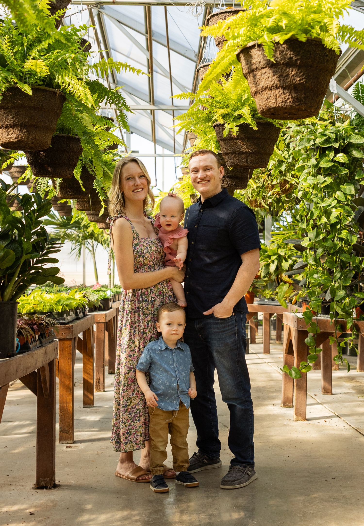 A family standing together in a greenhouse walkway with hanging ferns and wooden benches.