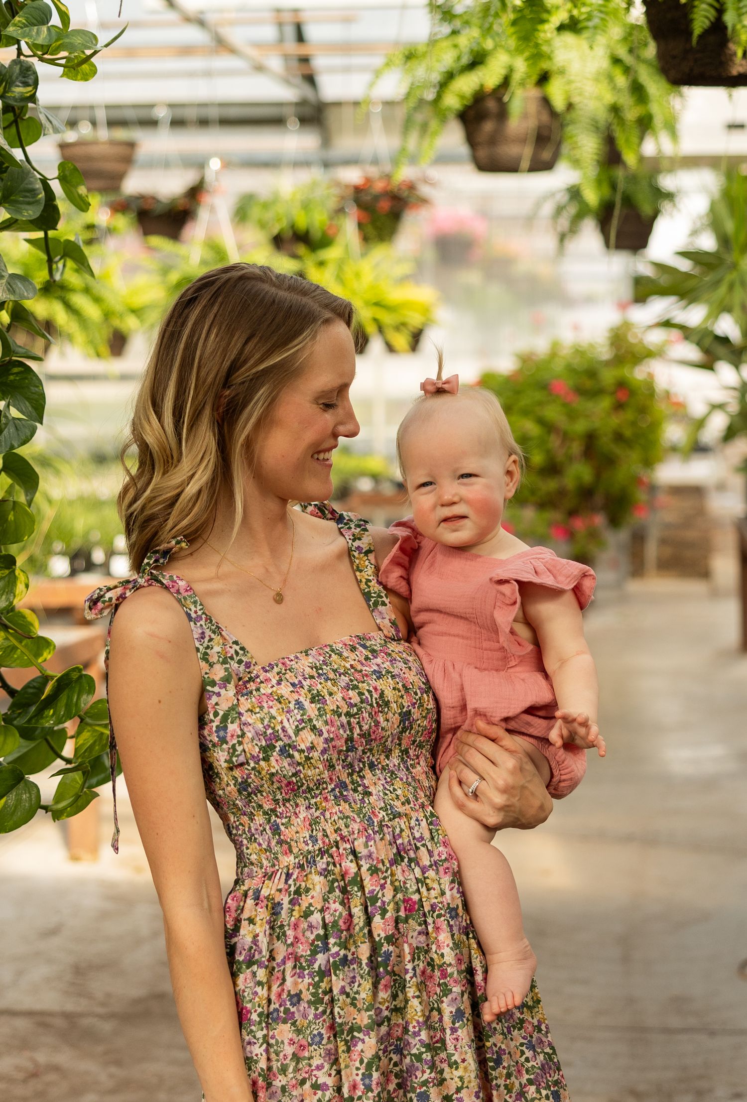 Woman in floral dress holding baby in pink outfit surrounded by greenhouse plants.