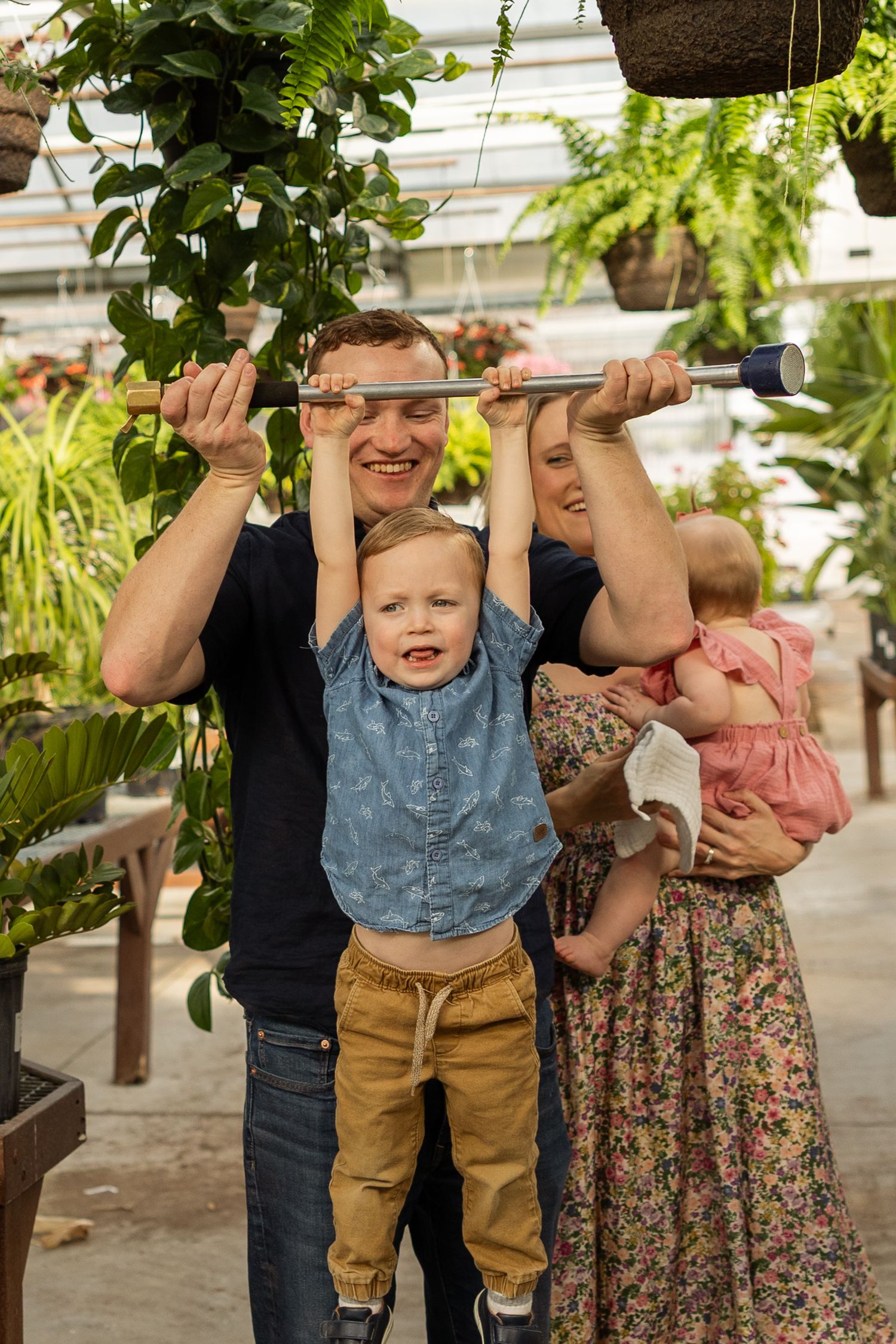 Family enjoying playful moments together in a greenhouse setting with hanging plants.