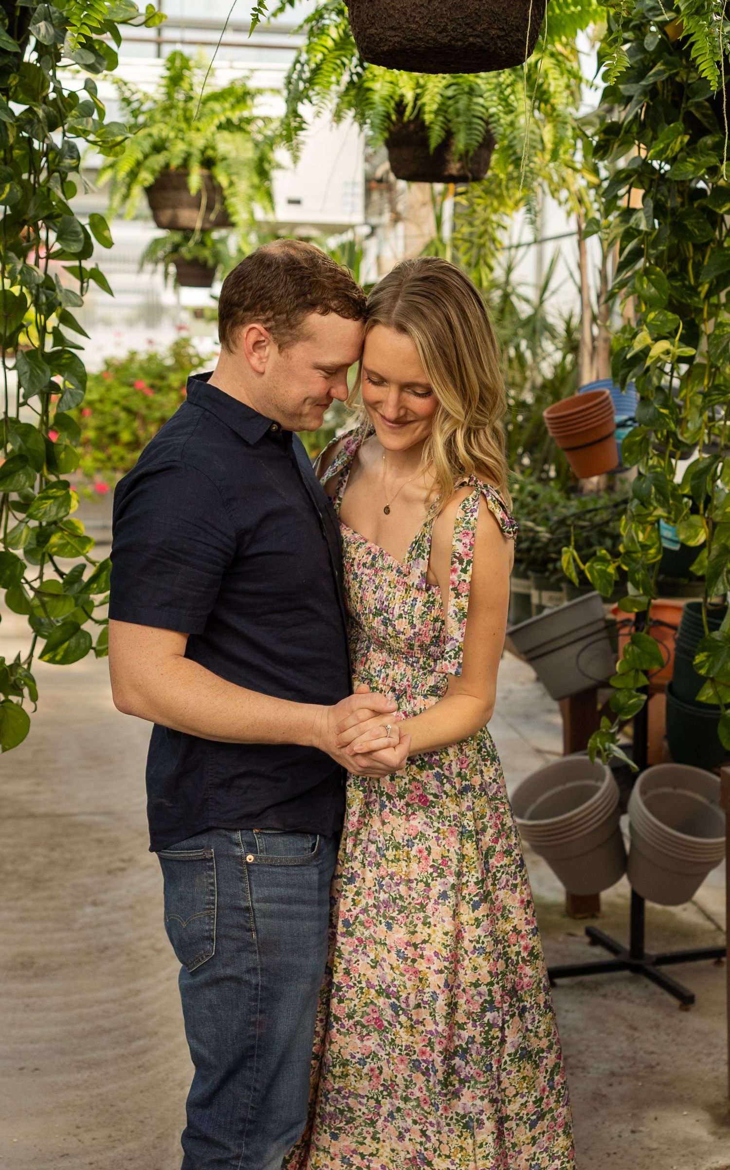 Romantic couple embraces while standing in a greenhouse filled with hanging plants.