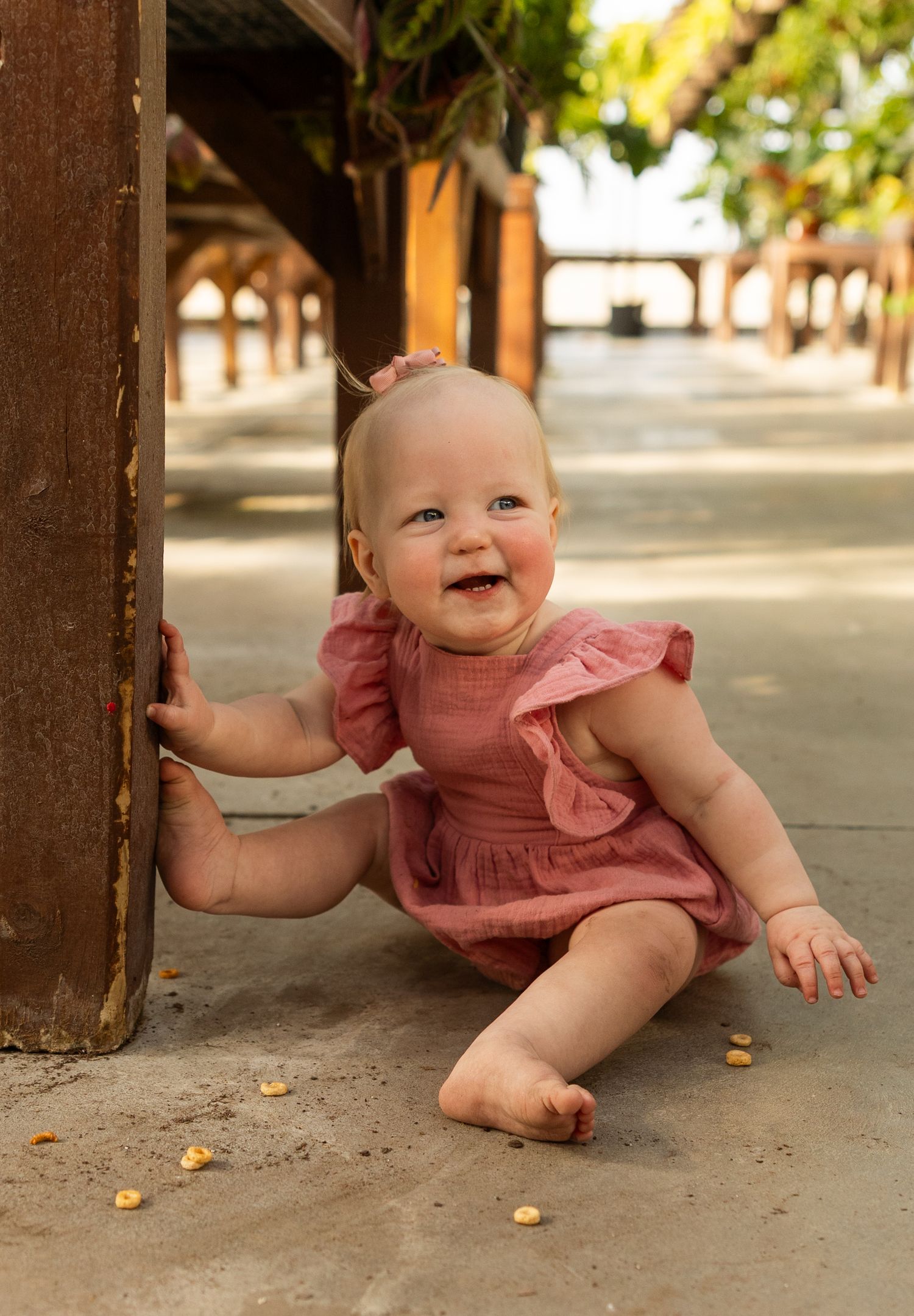 Baby in pink dress crawls and plays on concrete floor with scattered snacks.