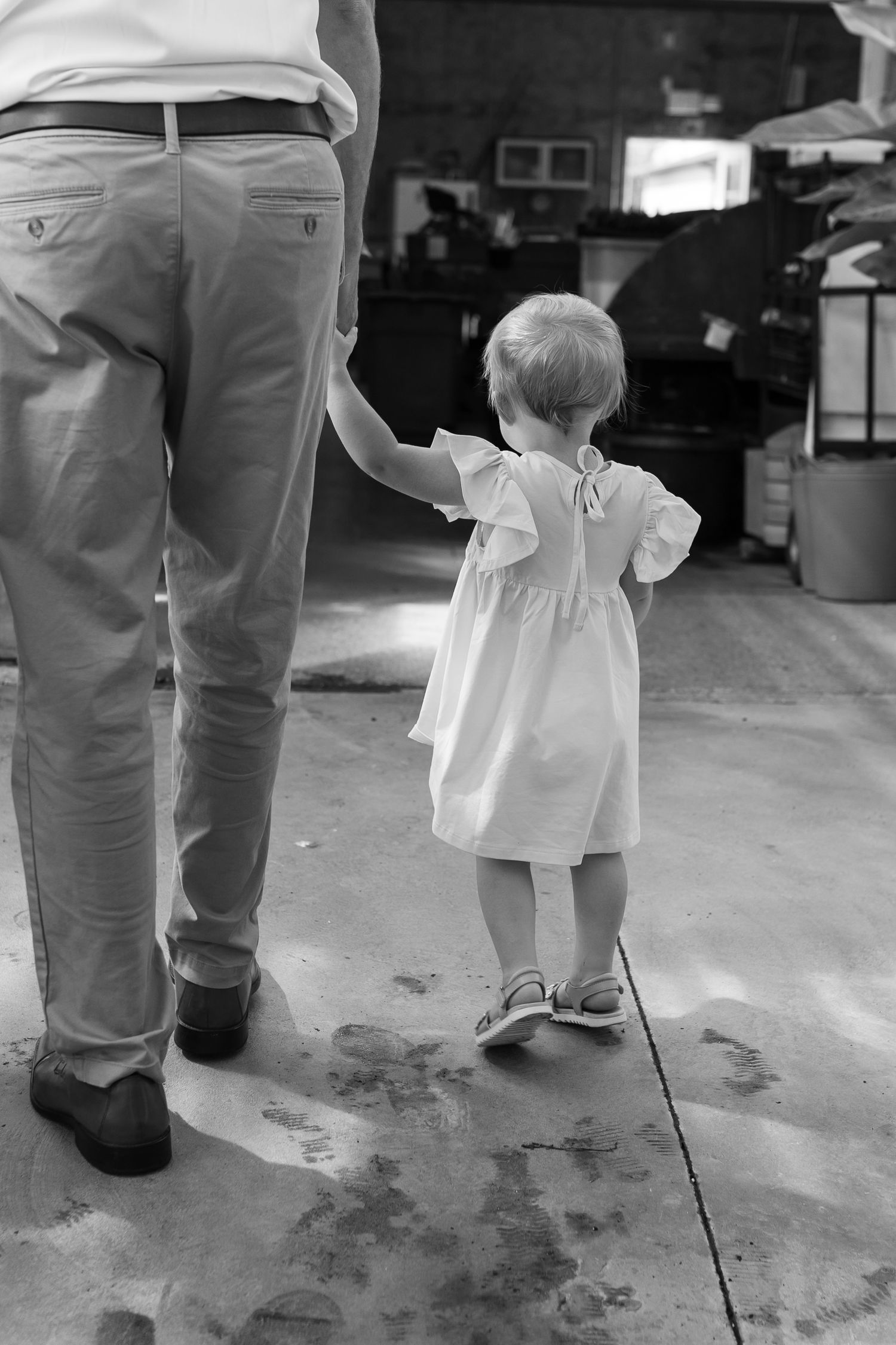 Black and white image of small child in white dress standing near adult legs.