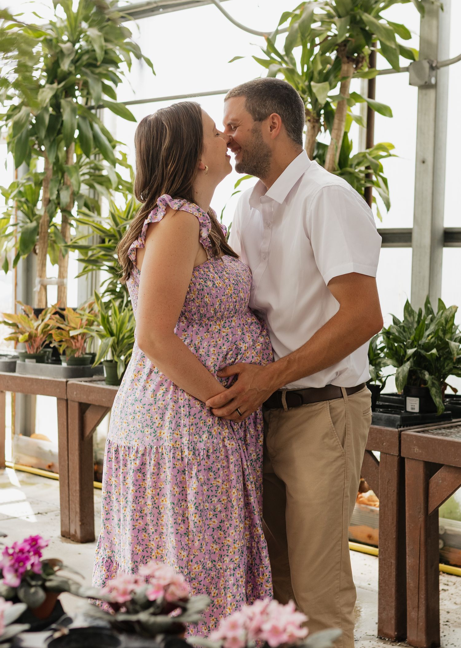 Intimate maternity photos in greenhouse with couple embracing among tropical plants.