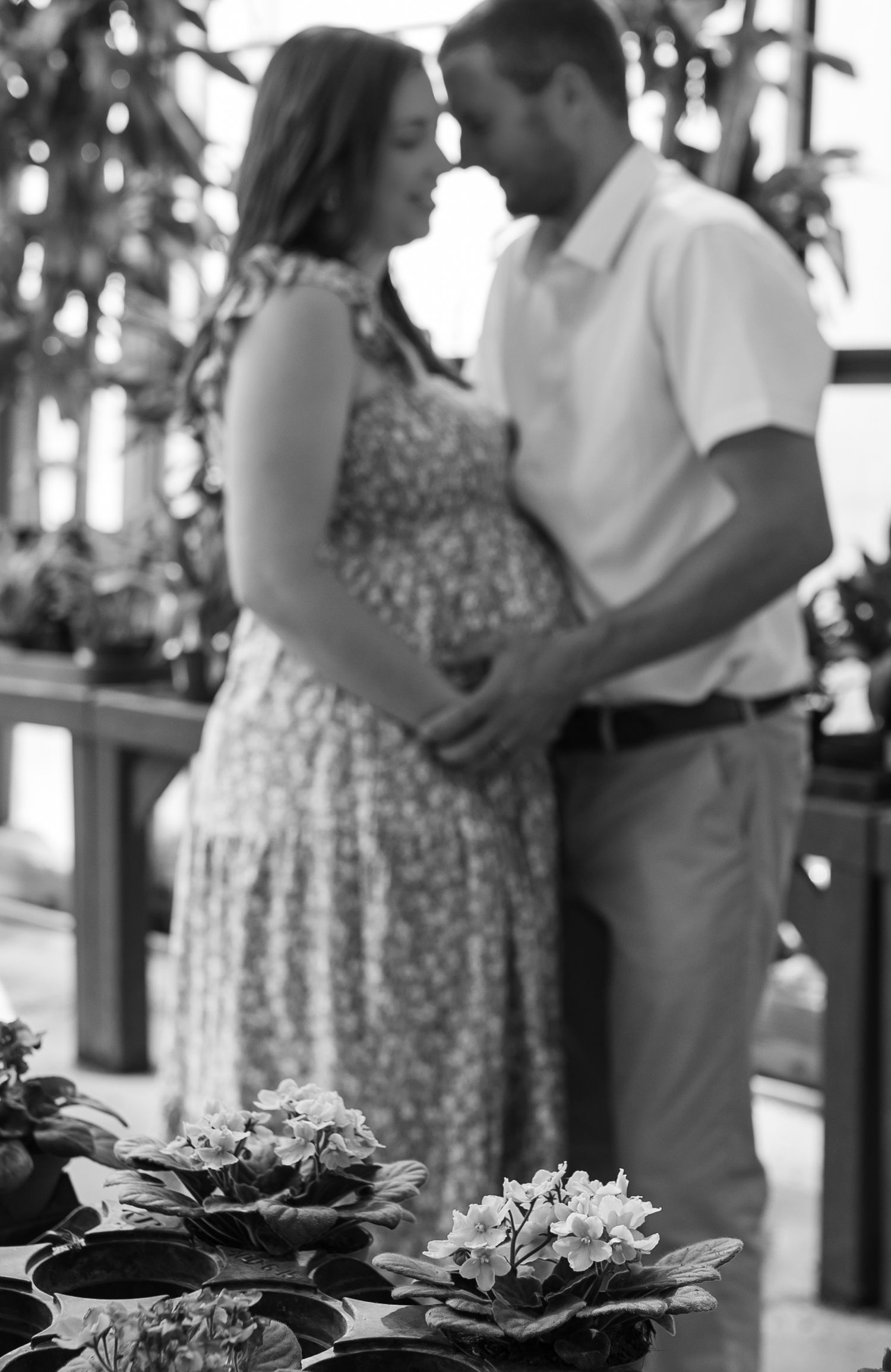 A romantic black and white photo of a couple embracing in a greenhouse setting with plants in the foreground.