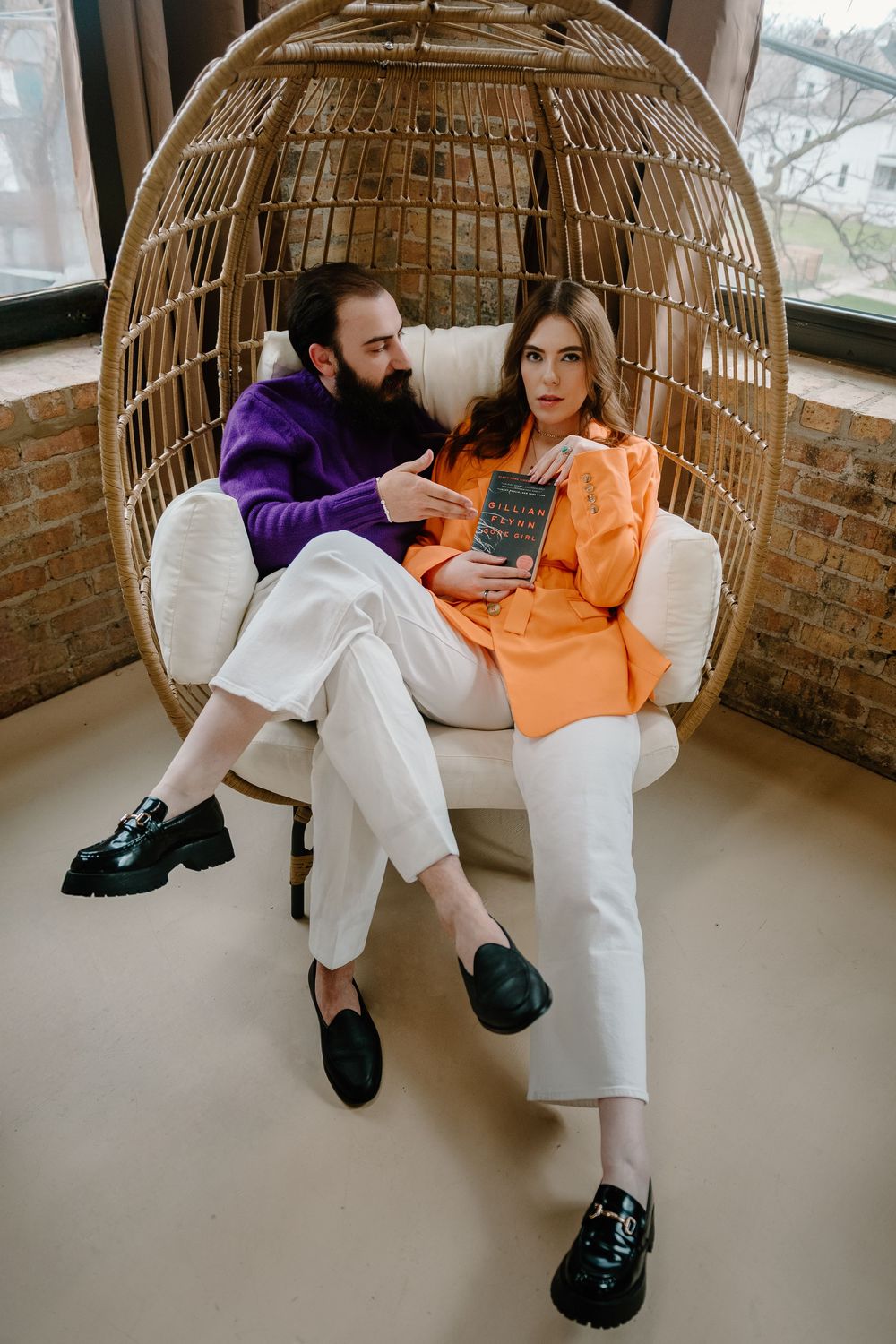 Two people lounging together in a modern hanging egg chair with orange and white outfits against a brick wall.