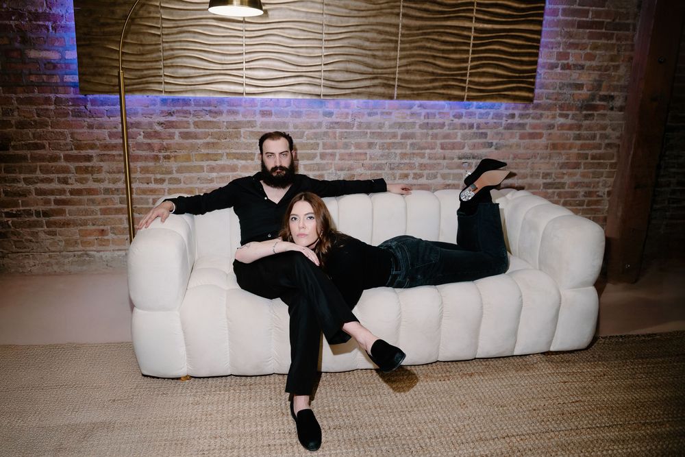 Two people lounging playfully on a white couch in a room with exposed brick walls and mood lighting.