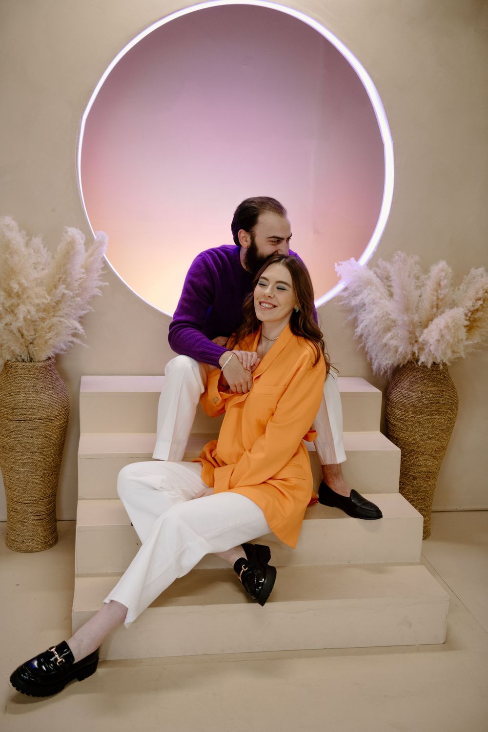 A couple poses on white steps against a circular pink backlight with pampas grass decor in woven baskets on either side.