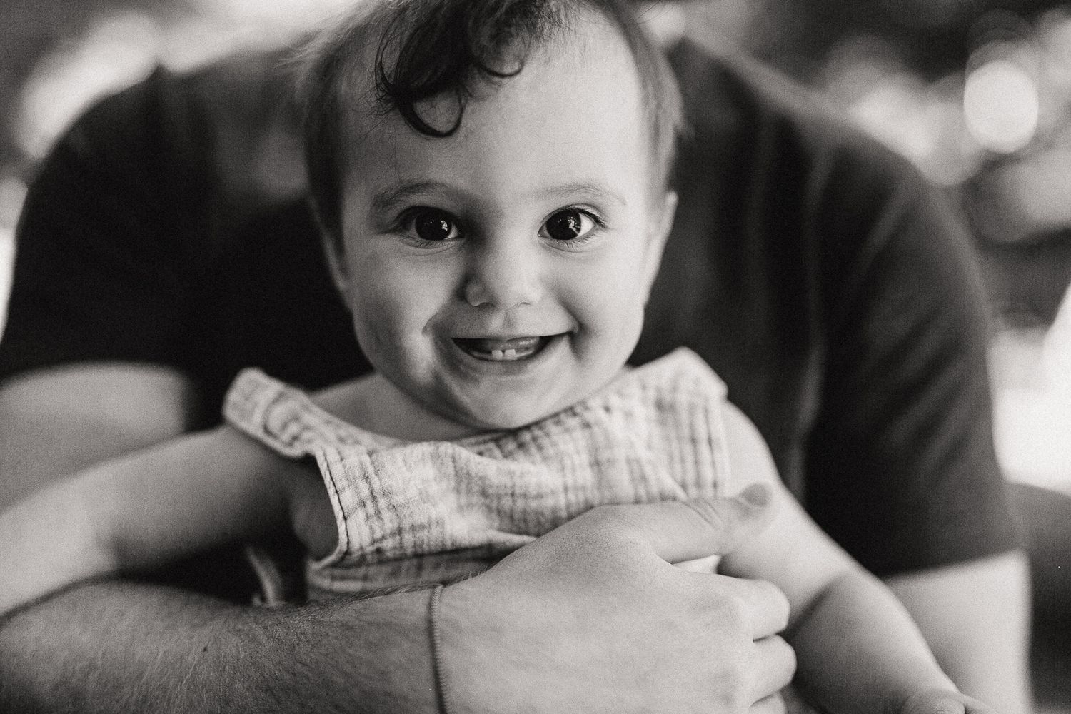 A black and white portrait of a smiling baby wearing a striped outfit while being held.