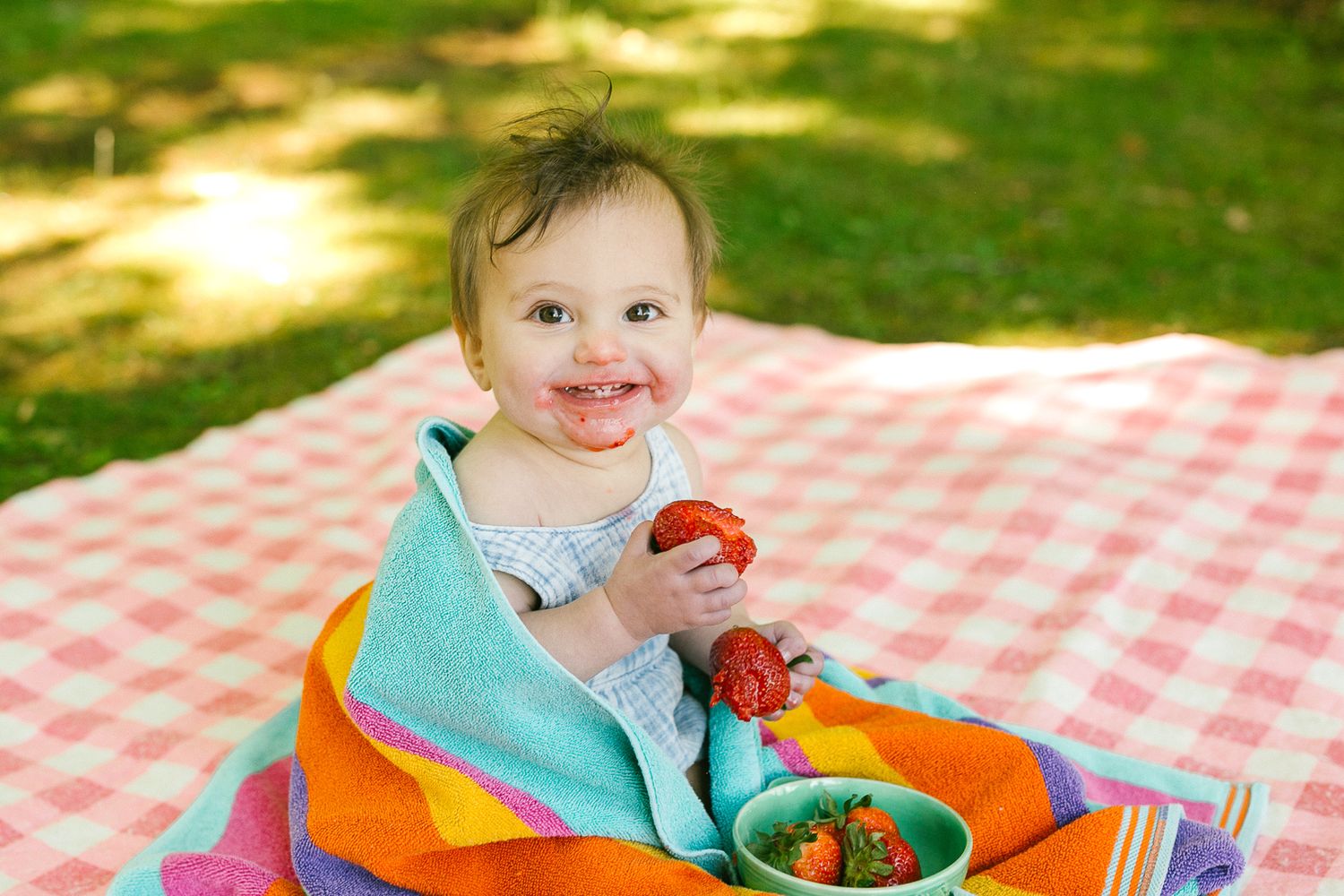 Child eating fresh strawberries at outdoor picnic on red and white checkered blanket on sunny day.