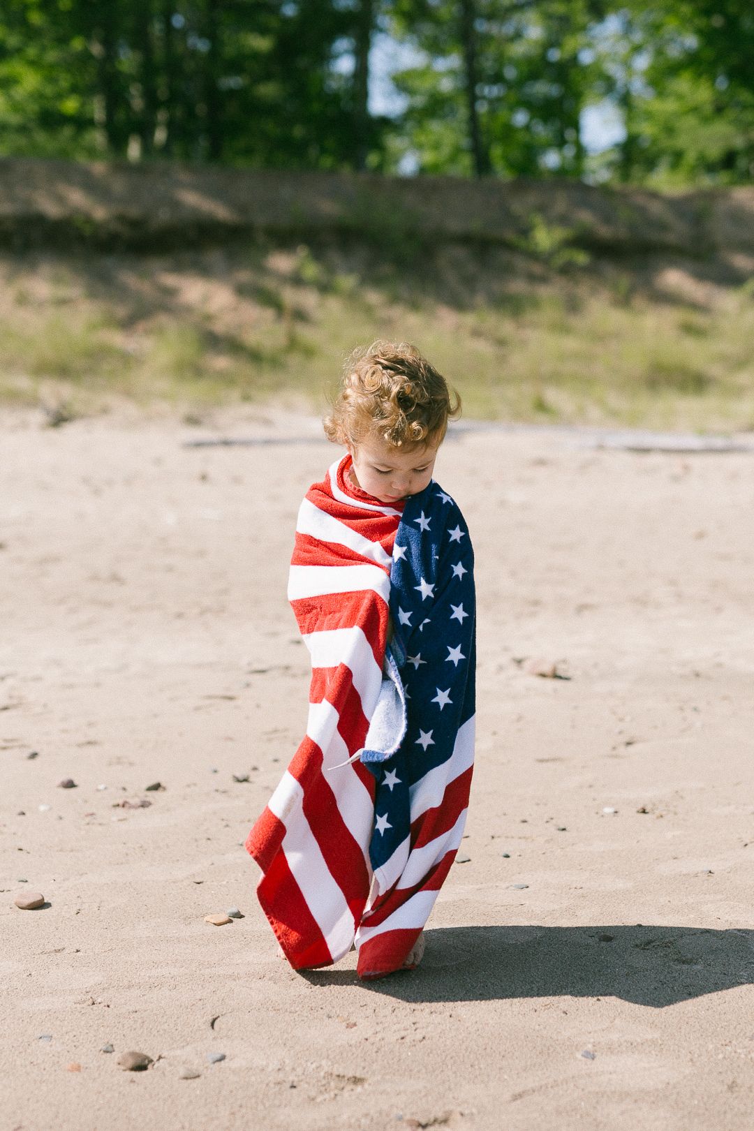 Small child wrapped in American flag blanket sitting on sandy beach on sunny day.