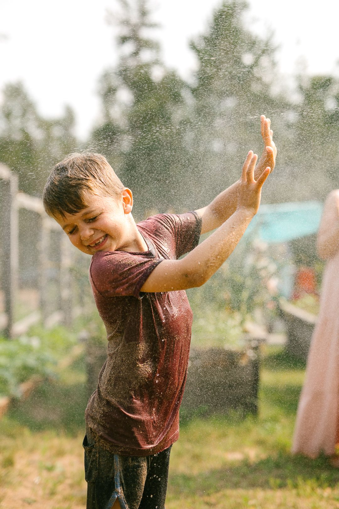 Child playing in water sprinklers on a sunny day in a garden with trees in background.