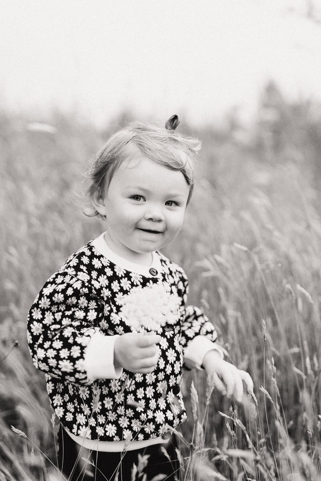 A toddler in a floral dress smiles while standing in a field of tall grass.