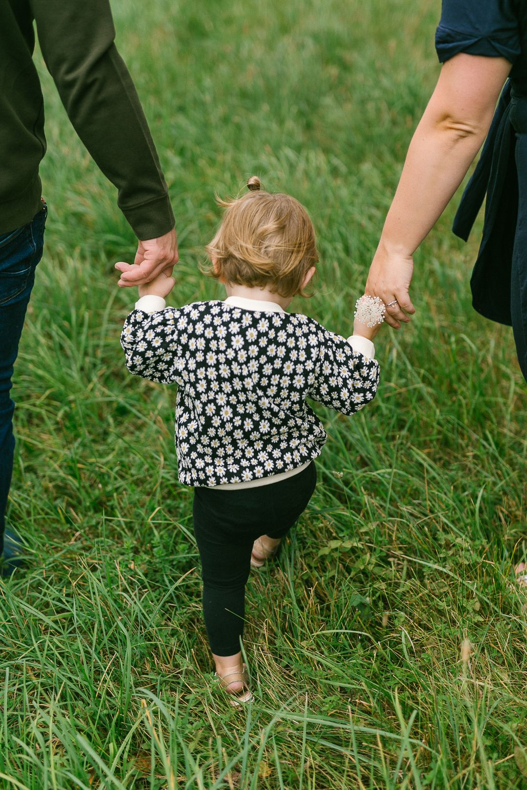 A small child in a floral print jacket holds hands with adults while walking through green grass.