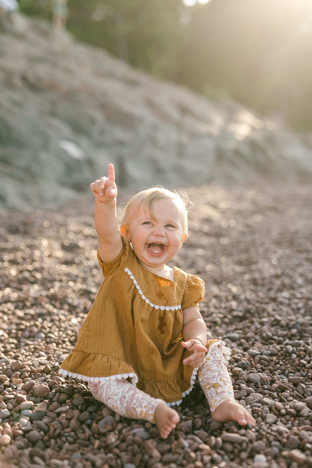 Happy baby in mustard dress sitting on rocky beach during golden hour.