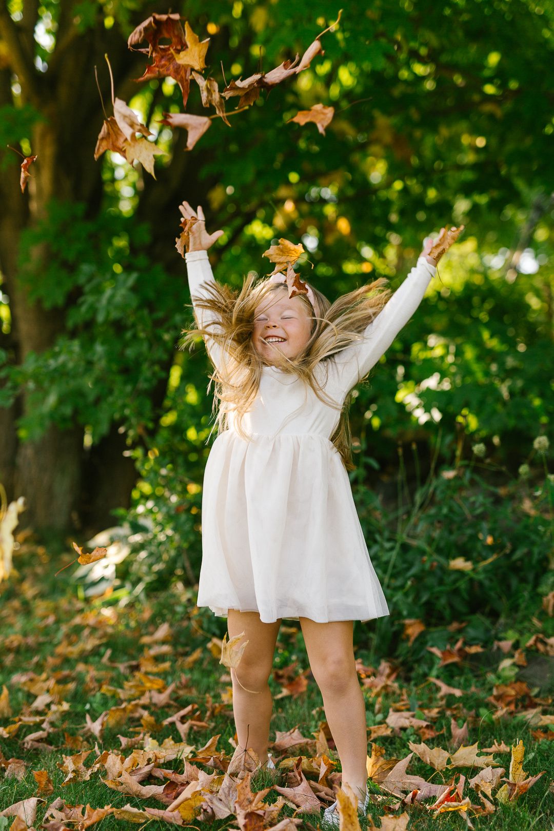 Young child in white dress playing with falling autumn leaves in a sunlit forest.