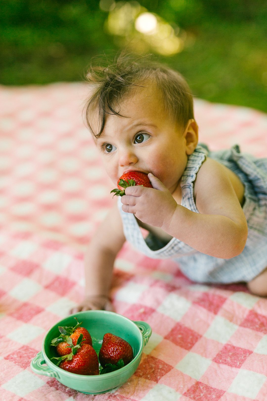 A child peeks playfully over the edge of a red and white checkered tablecloth at a bowl of fresh strawberries.