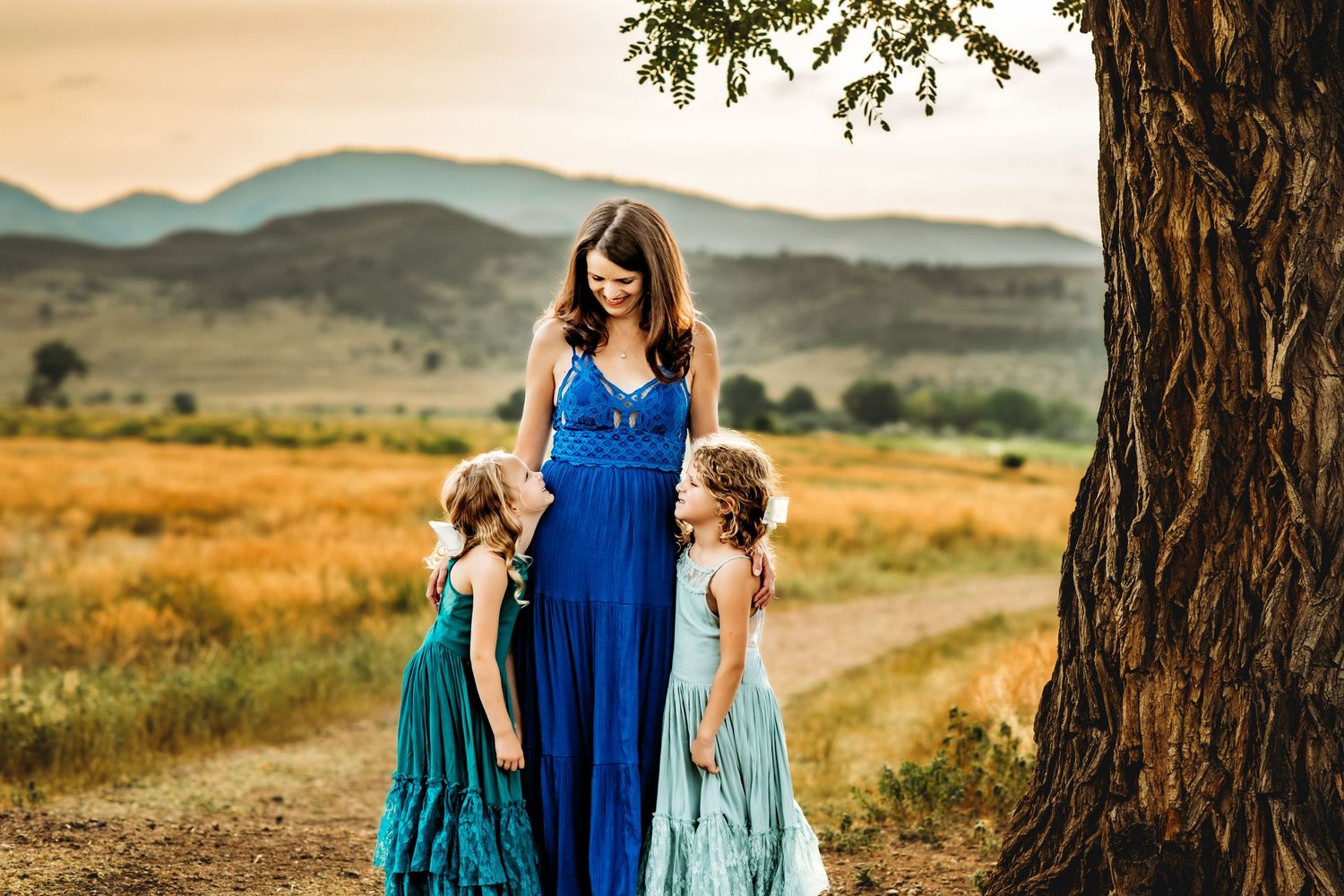 Family in coordinating blue dresses poses together in golden field with mountains in background at sunset.