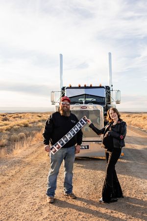 Two people stand in a desert landscape with trucks and equipment on a dirt road at sunset.