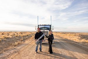 A panoramic view of a desert road stretches into the distance with people and vehicles visible on the horizon.