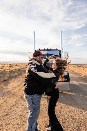 Two people embrace on a desert road with dramatic clouds and arid landscape in the background.