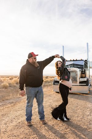 Two people dancing together on a dirt road with a semi truck parked in the desert landscape behind them.