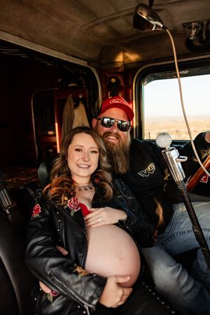 A couple in leather jackets sits together inside a vehicle, with one wearing a red beanie and sunglasses.