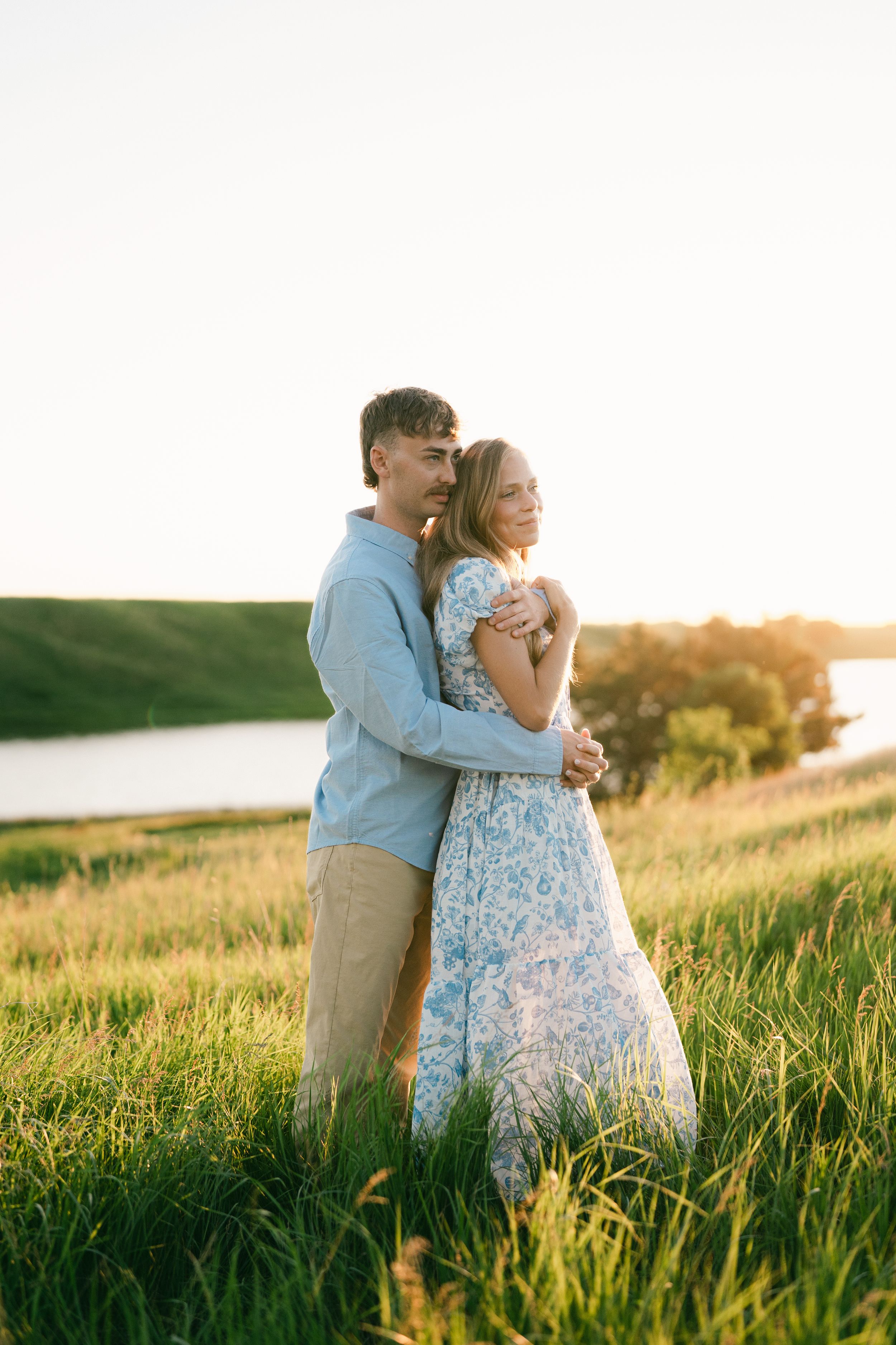 Couple embracing in golden hour sunlight on green hillside near lake, woman in floral dress, man in blue shirt.