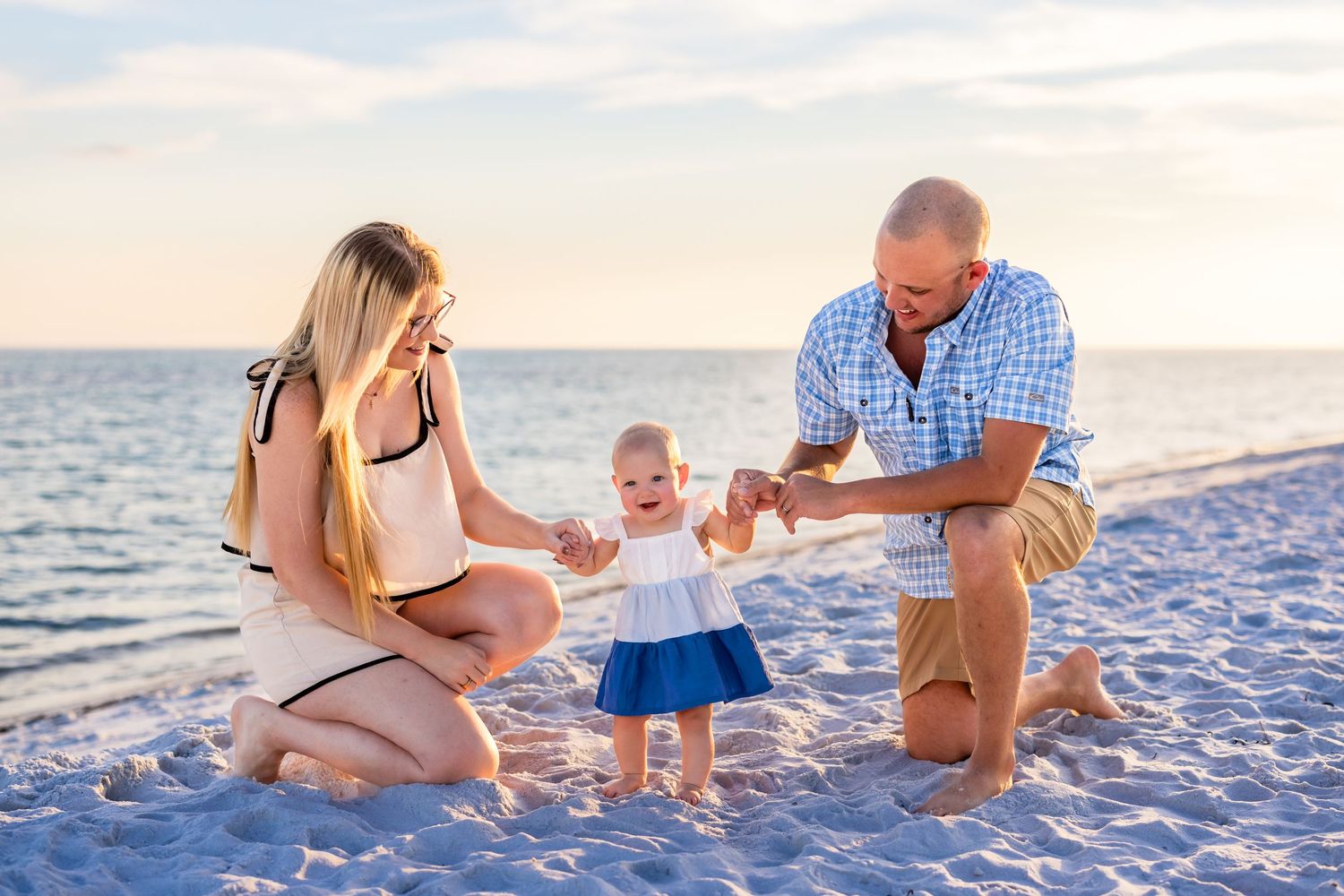 Family enjoying a sunset beach photoshoot with their baby in matching white and blue outfits.