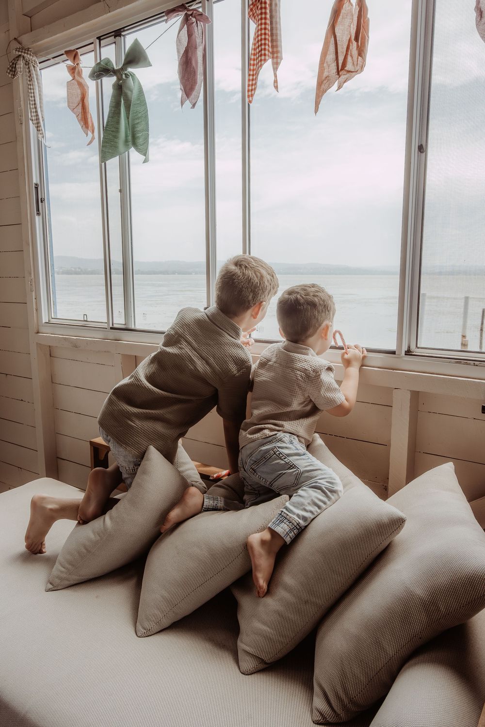 Children lounge on cushions by large windows overlooking a coastal view in a cozy sunroom at a central coast christmas photoshoot.