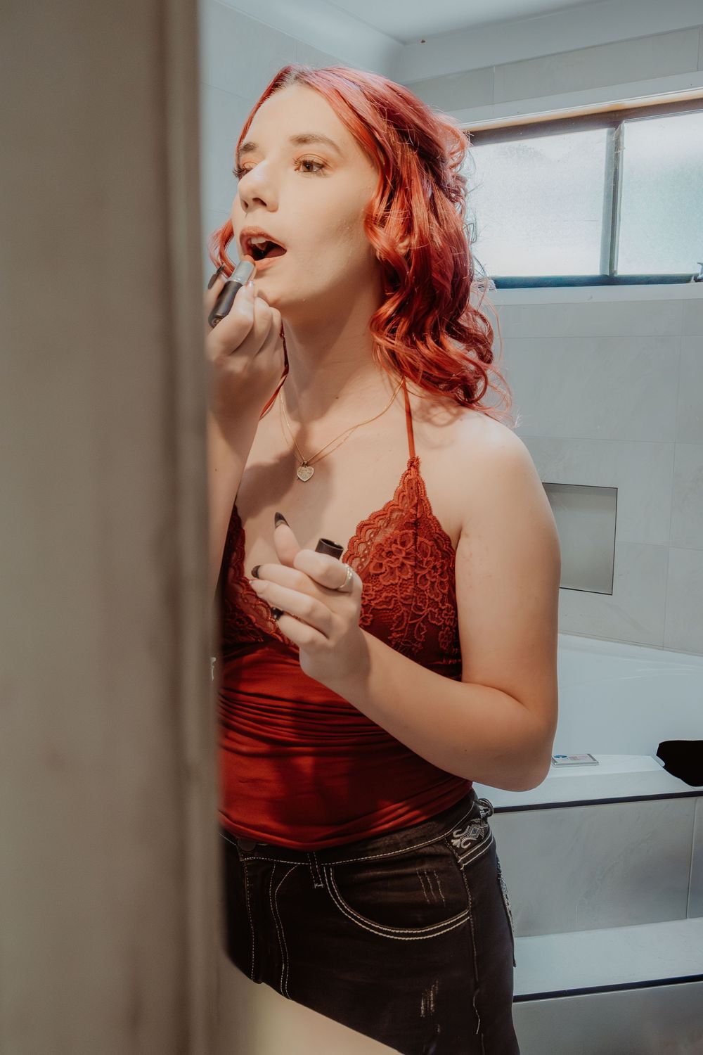 Person with bright red hair applying makeup in front of bathroom mirror wearing burgundy top.