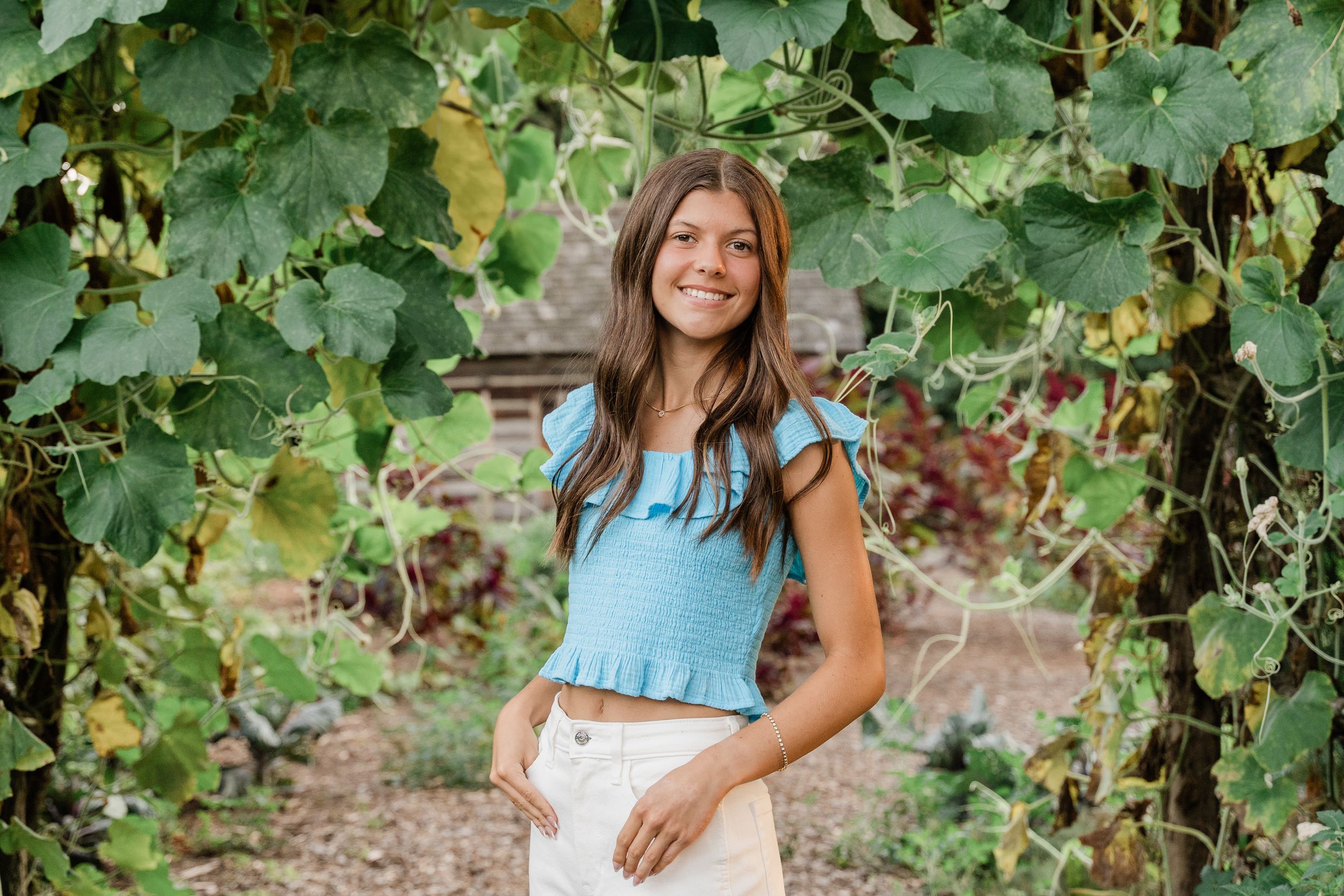 A person in a blue dress poses among lush grape vines in a sunny vineyard setting.