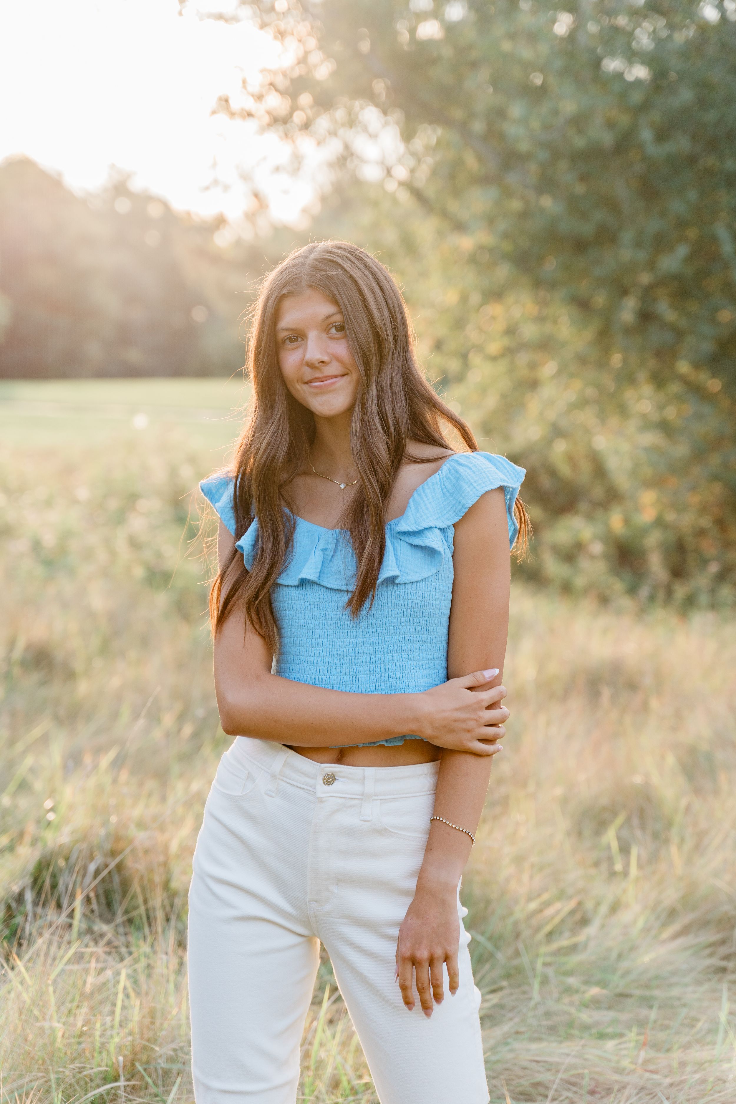 A person in a light blue top and white pants poses outdoors in a sunlit field during golden hour.