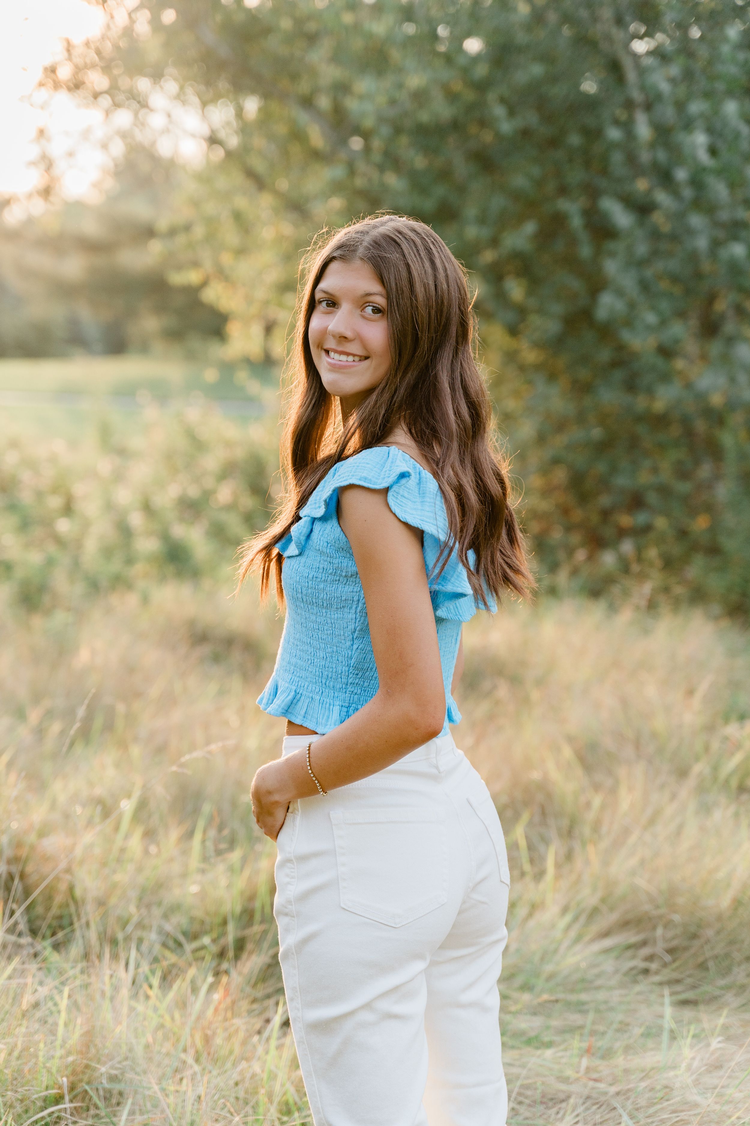 A maternity photoshoot in a sun-drenched field shows a woman in a blue top and white dress turning playfully.