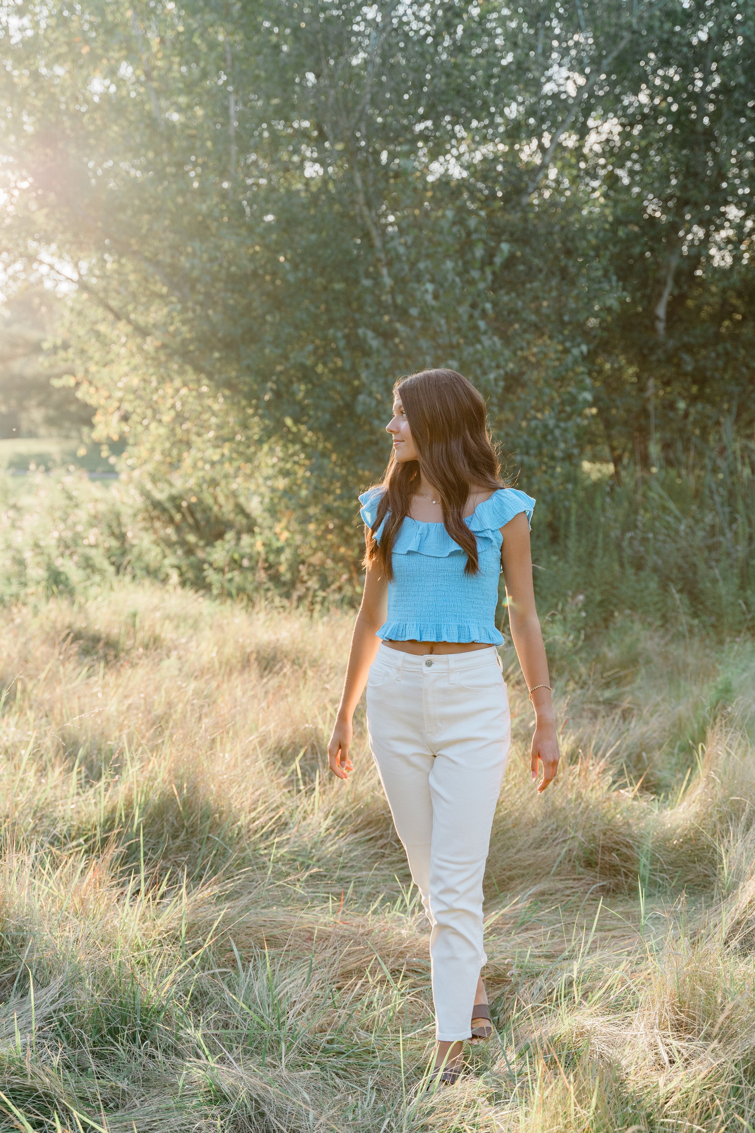 Person in light blue ruffled top and white pants walking through sunlit meadow with trees in background.