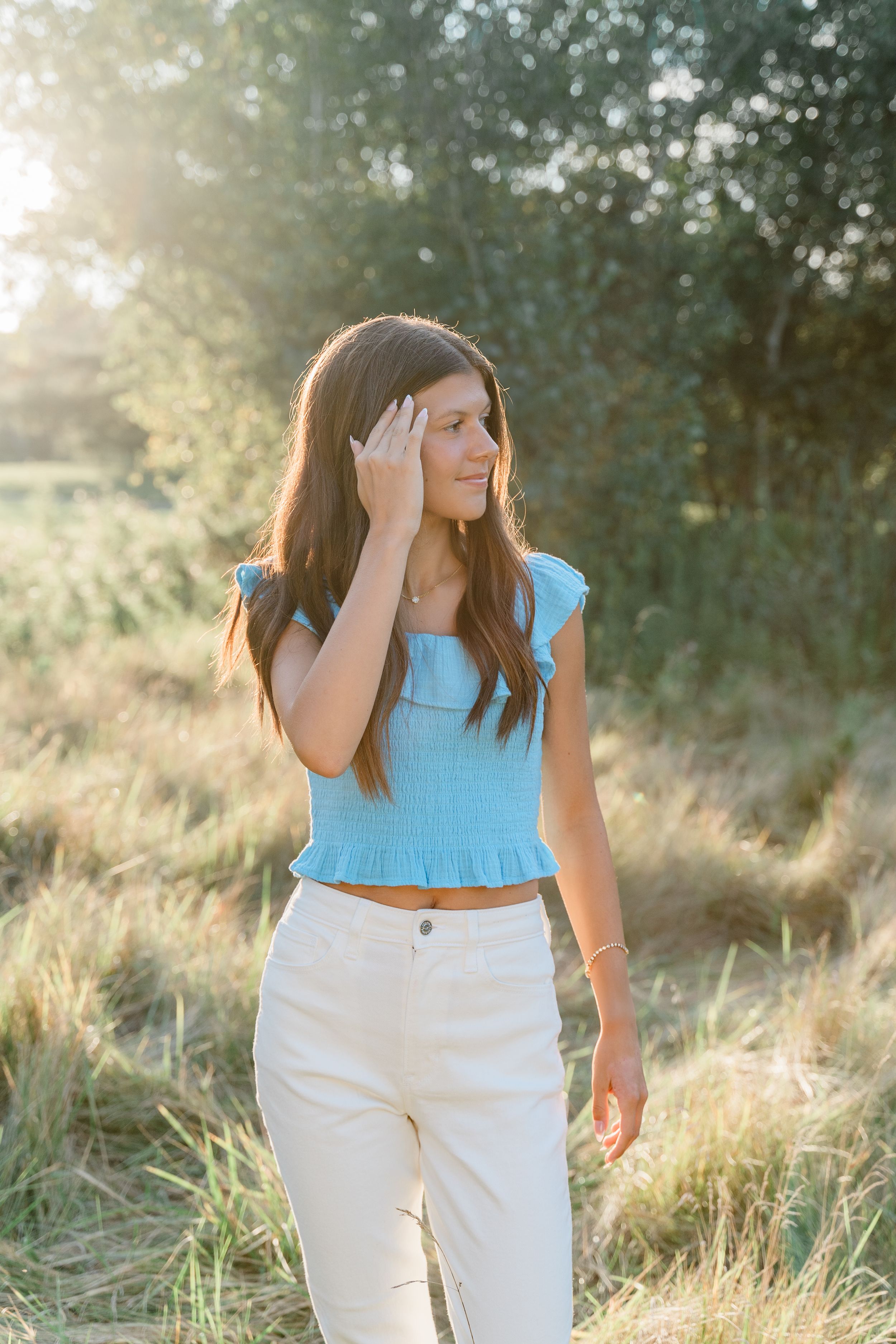 Woman in light blue top and white pants poses in golden sunlit field during sunset.