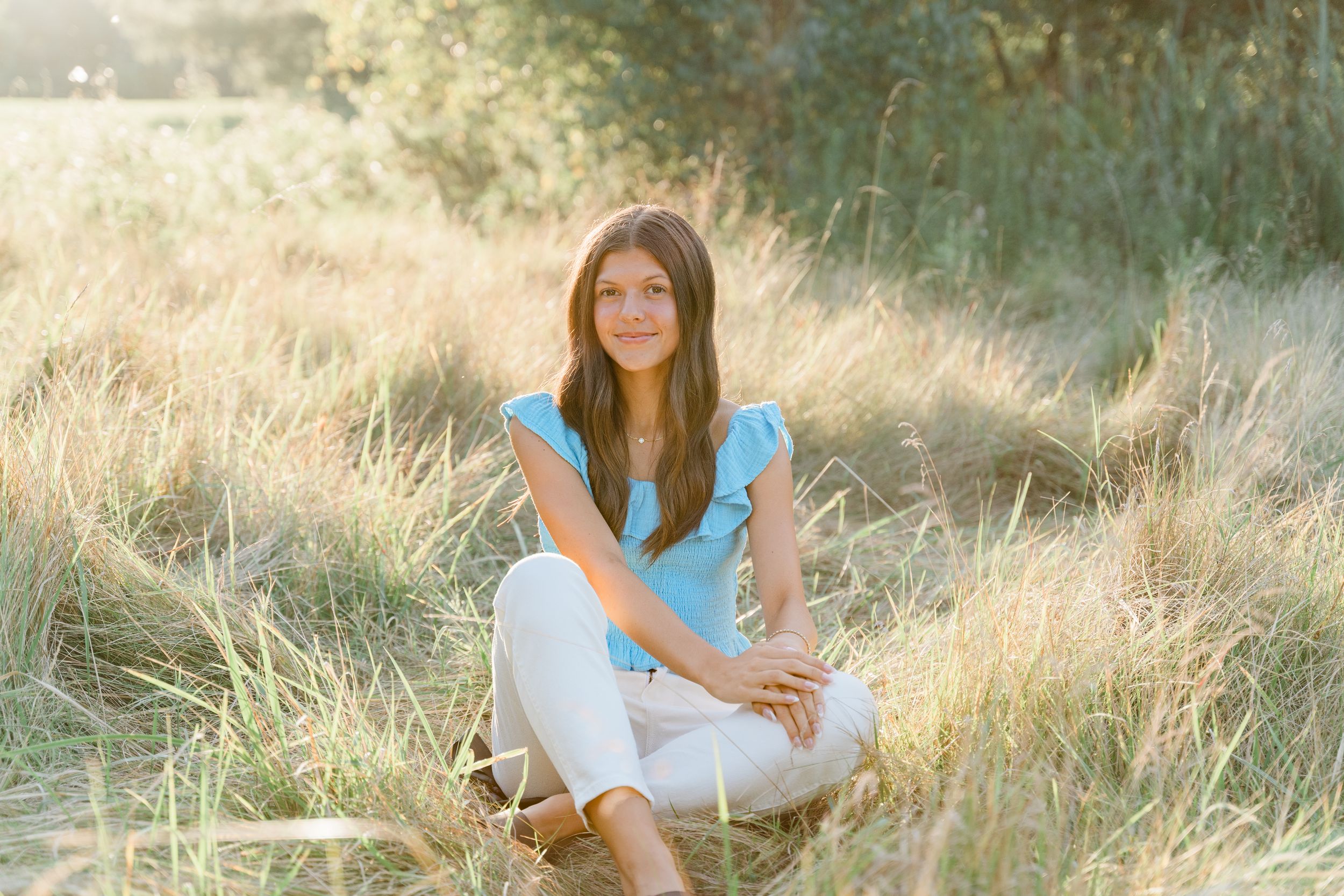 A person in a white dress sits gracefully in a sunlit meadow with tall golden grass and natural lighting.