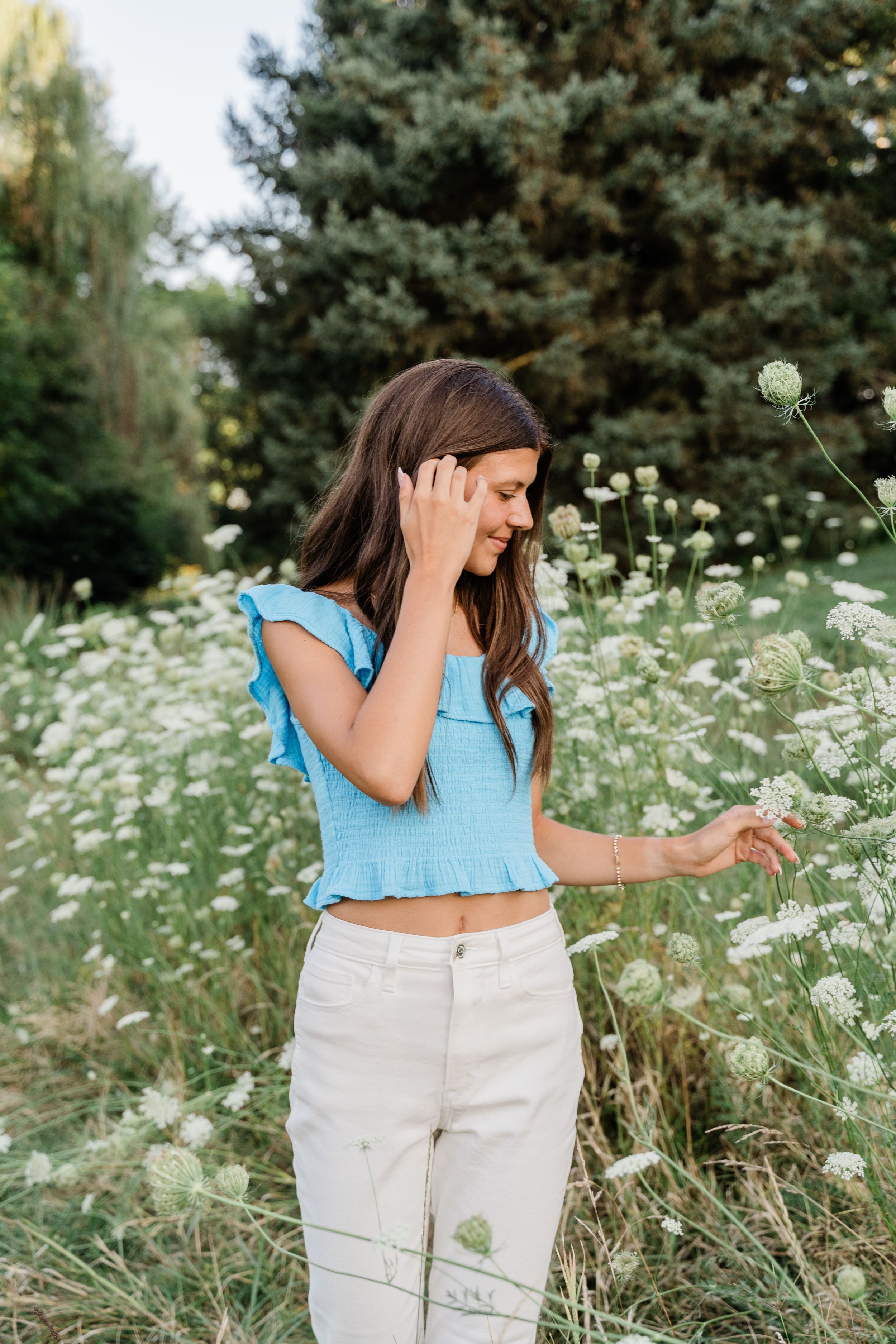 A person in white pants and blue top stands in a field of blooming white daisies on a sunny day.