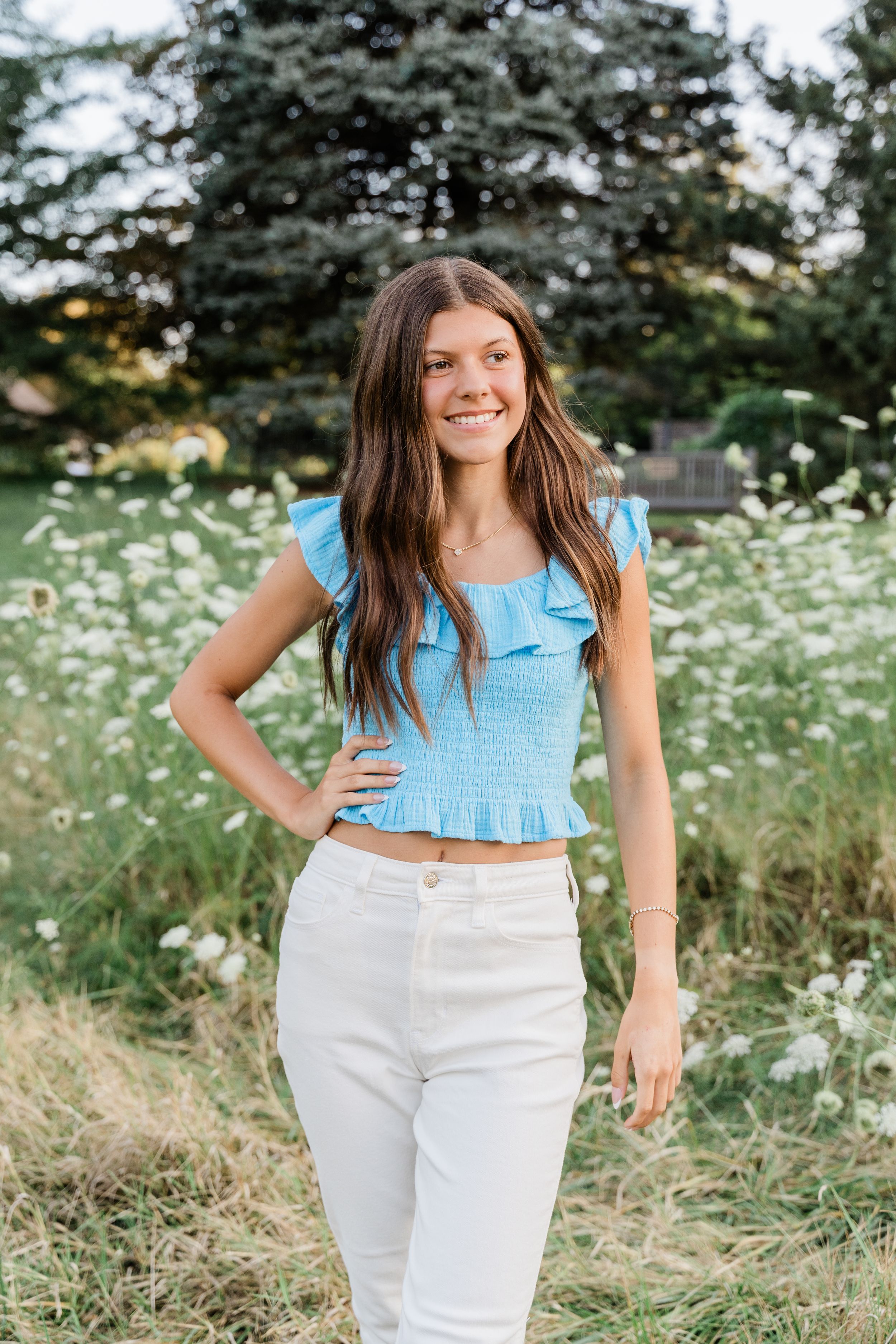 A person wearing a light blue crop top and white pants poses in a field of white wildflowers and tall grass.