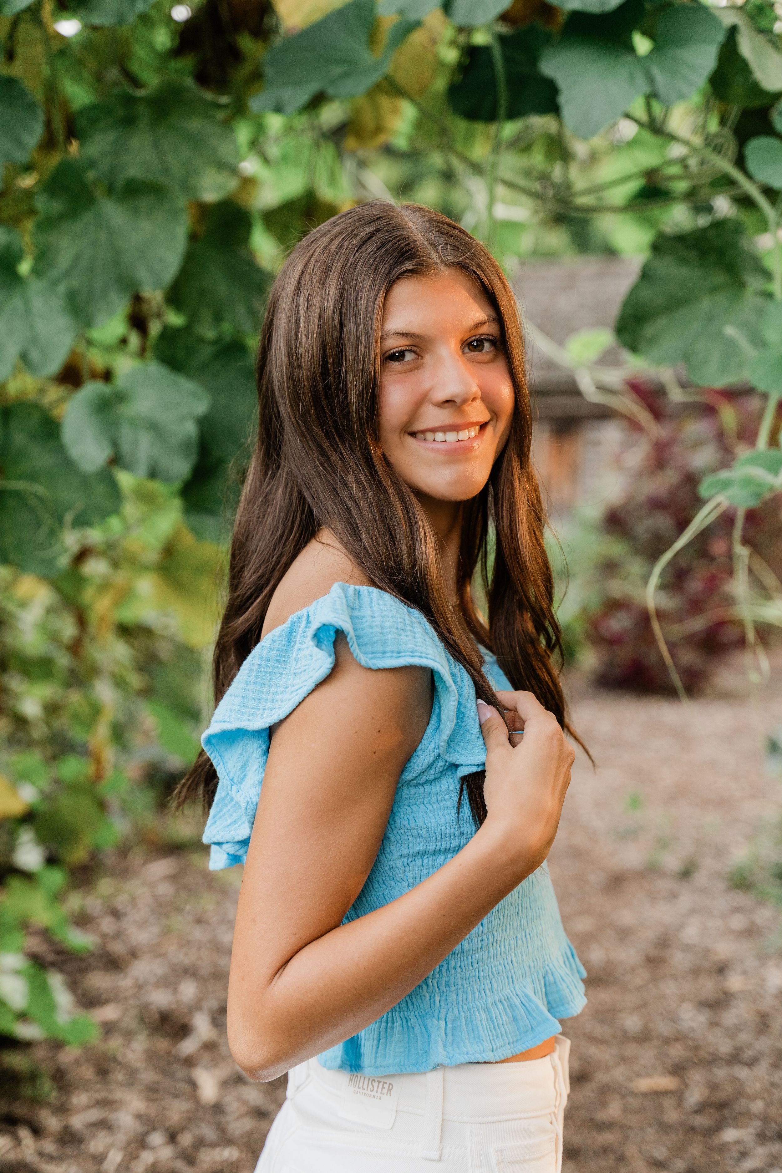 A person in a light blue ruffled crop top poses against a leafy green background in summer.