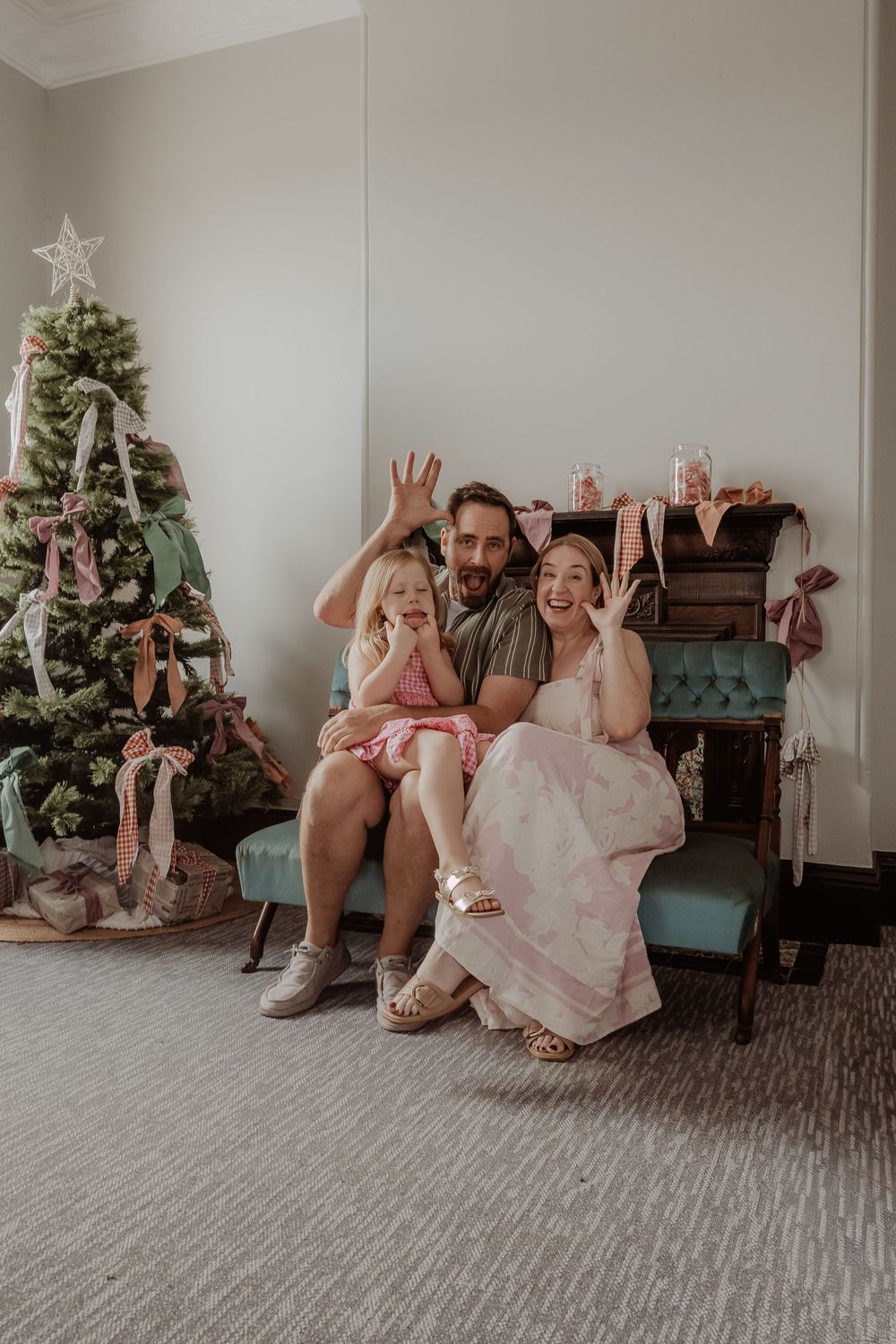 A family sitting on a teal blue couch playing peek-a-boo with a Christmas tree in background at a hunter valley christmas photoshoot.