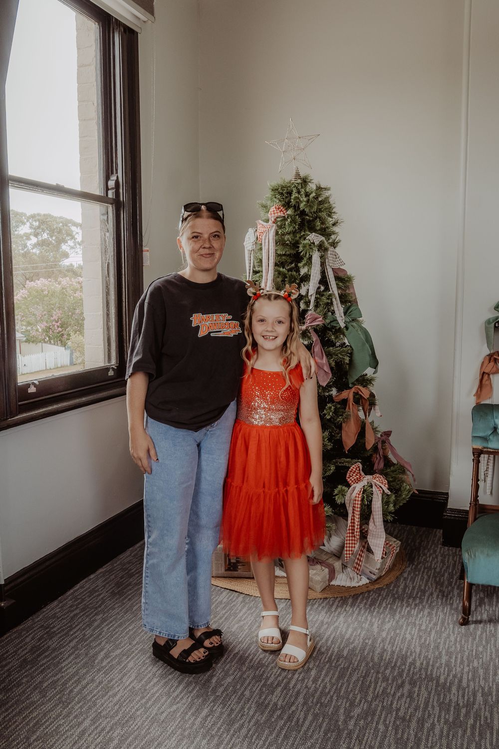 Two people stand together in a bright room with large windows and gray carpeted floors at a hunter valley christmas photoshoot.