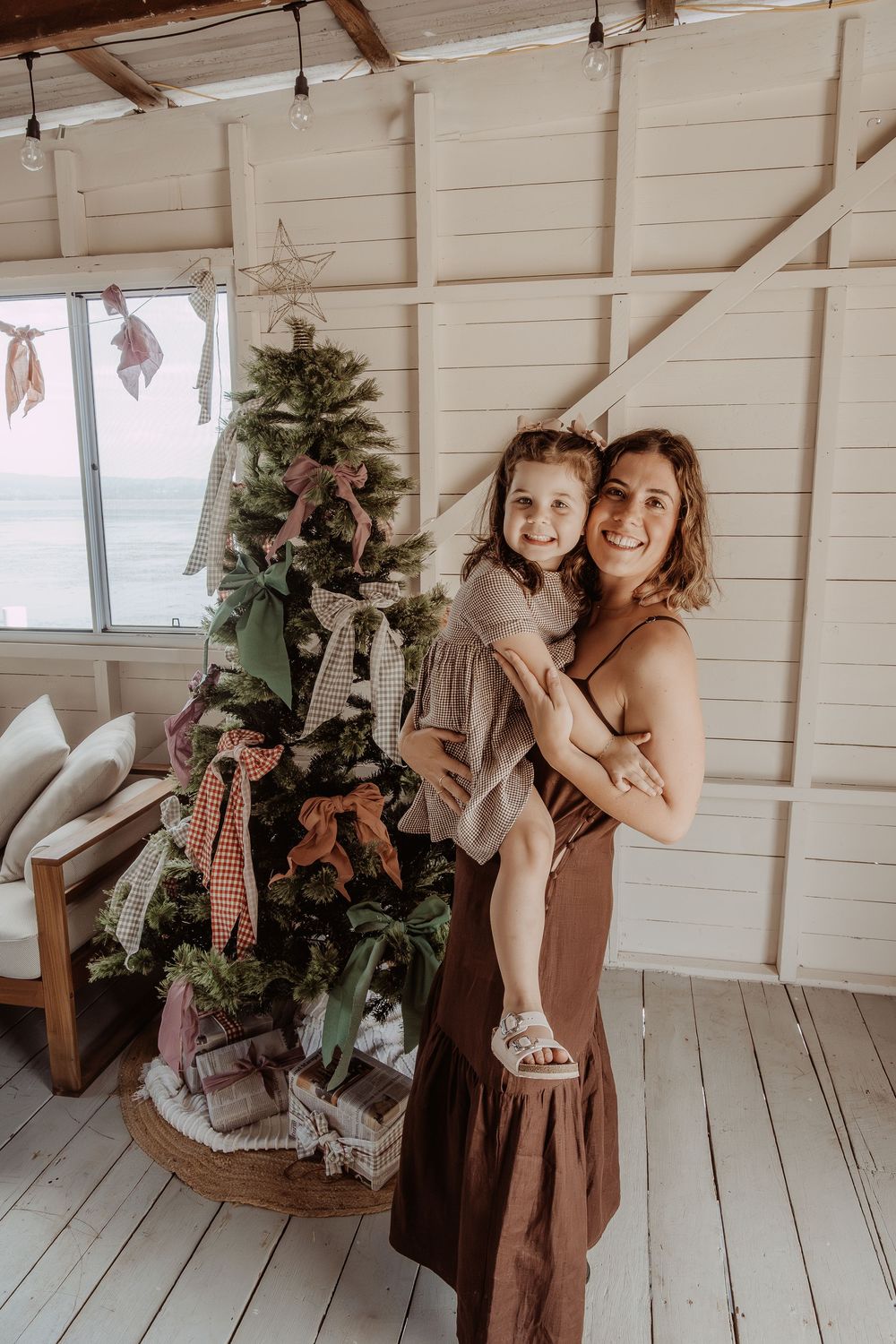 Two people in brown dresses laughing and embracing in a rustic white wooden room at a central coast christmas photoshoot.