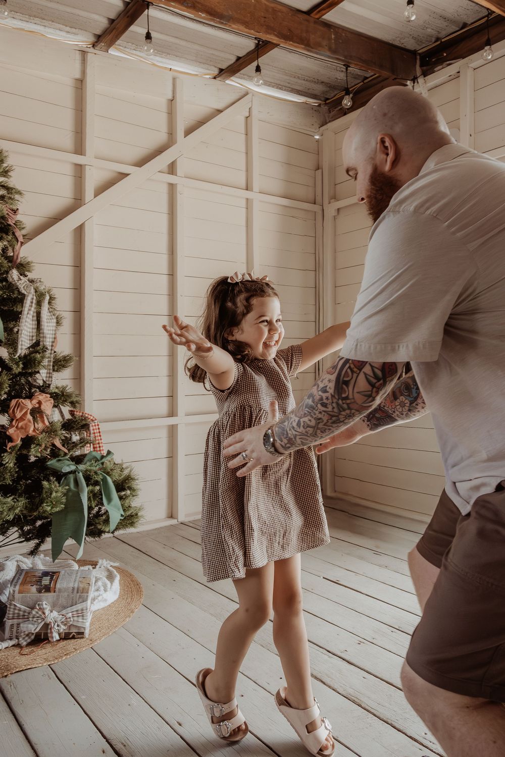 A young child in a dress dances with her dad in a white t-shirt next to a decorated Christmas tree at a central coast christmas photoshoot.