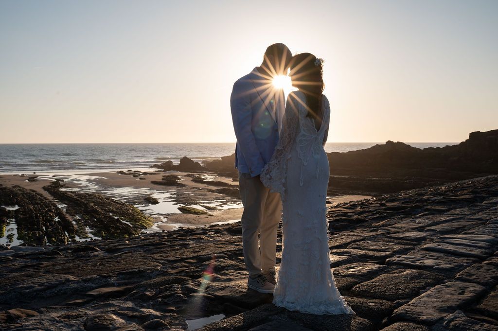 Bride and groom stand close together on the Bude breakwater at the end of the beach. They setting sun shines through between them.