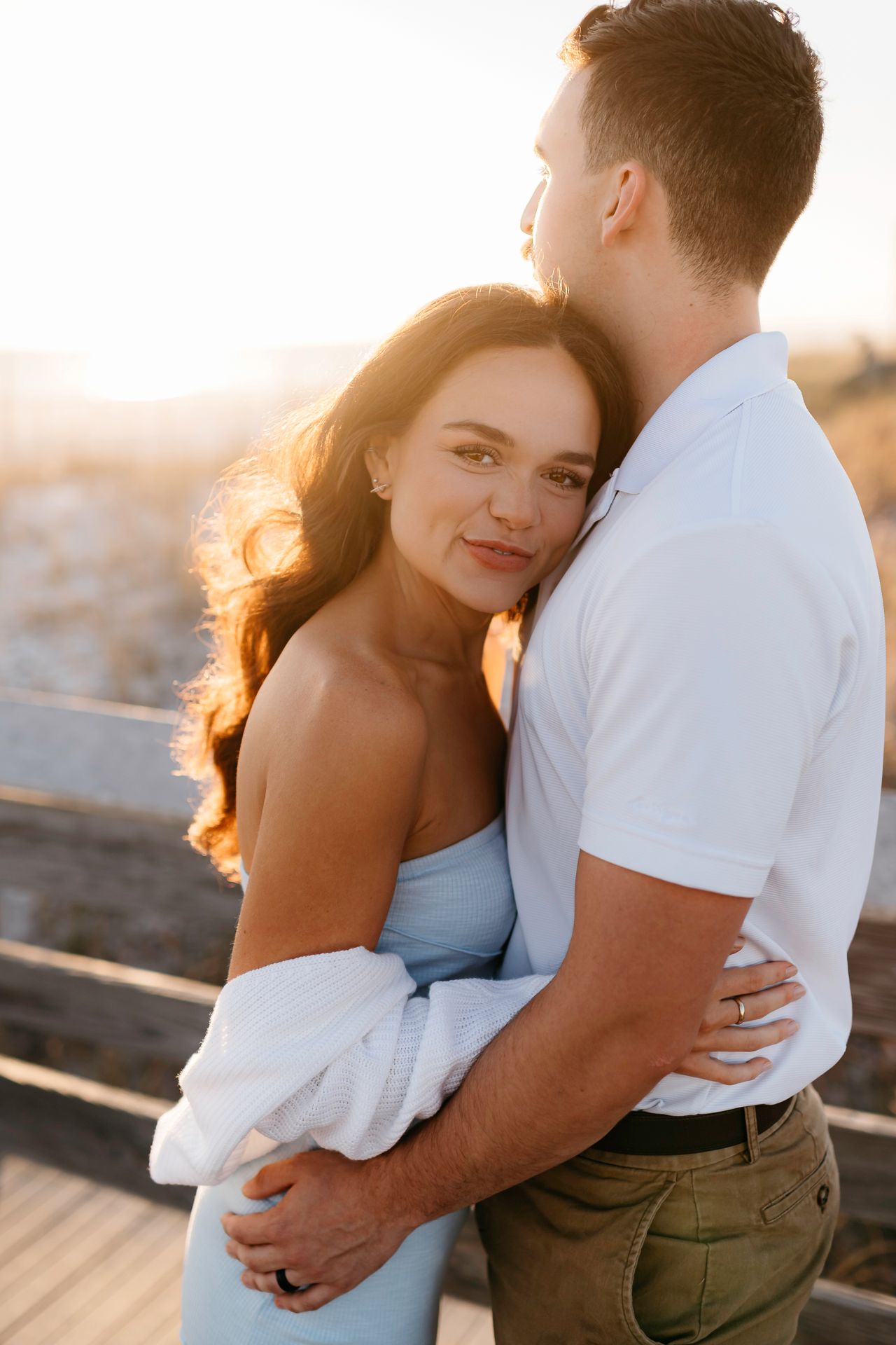 A couple embraces during golden hour on a deck, backlit by warm sunlight creating a romantic silhouette.