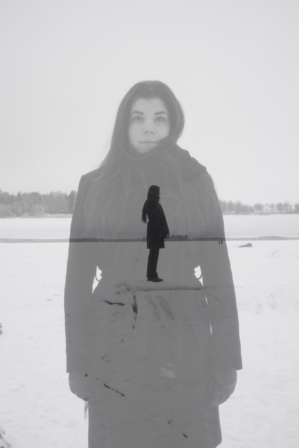 A person stands in a snowy winter landscape wearing dark clothing against a stark white background.
